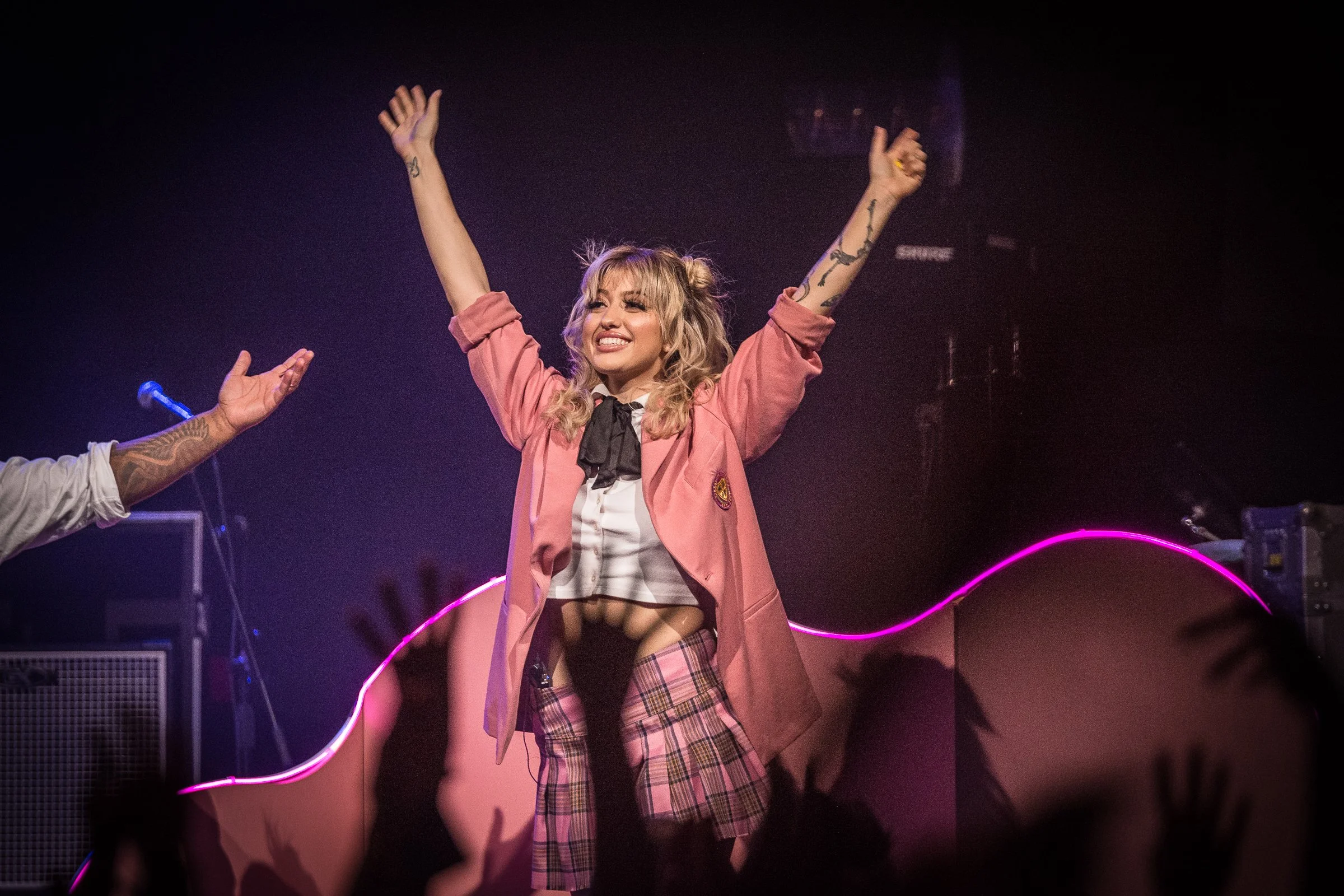 A woman on stage with arms raised, smiling, wearing a pink jacket, tartan skirt, and black stockings, during a performance or event in a dark, lit-up setting.