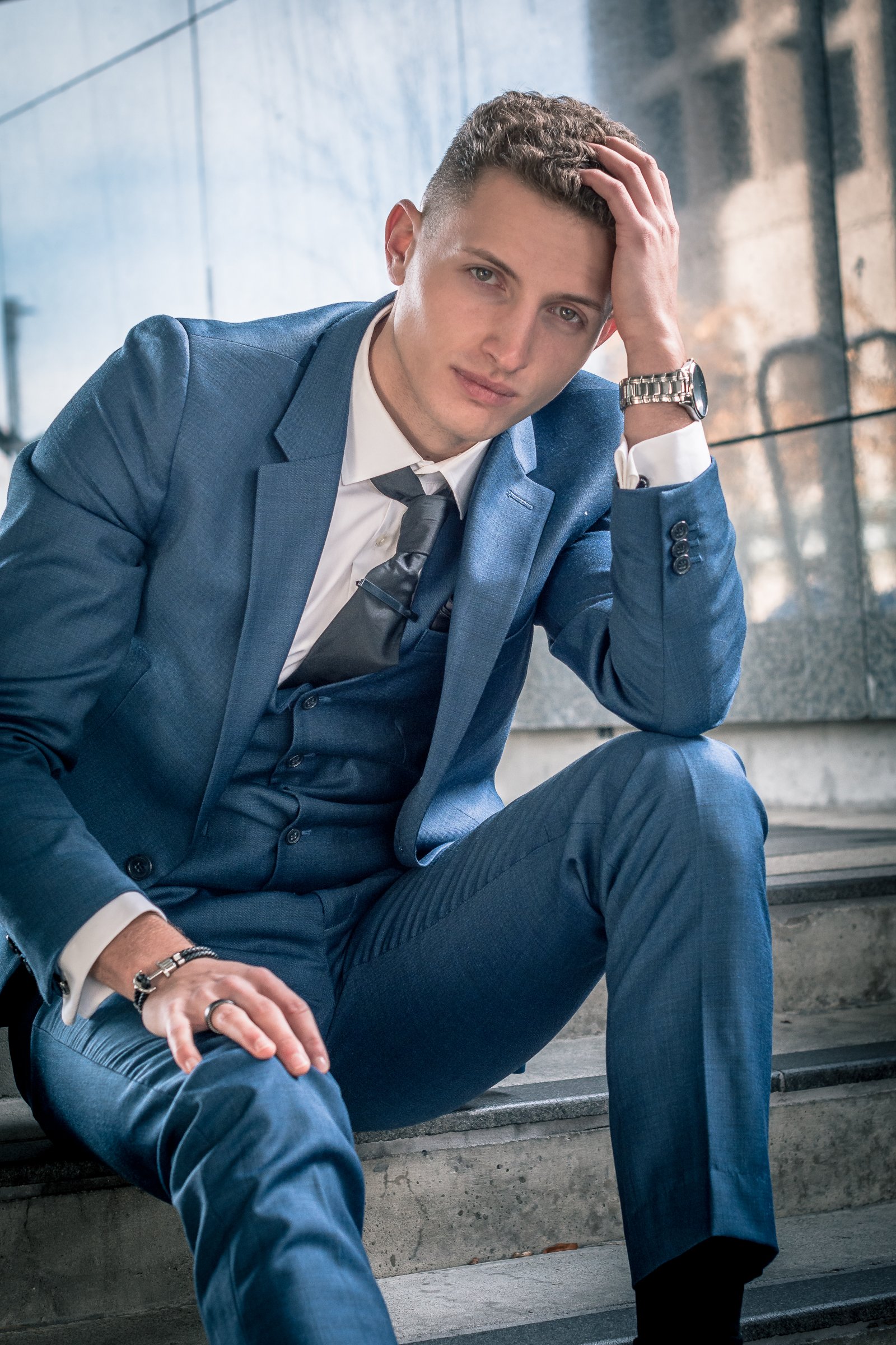 A young man wearing a blue suit, white shirt, and dark tie sitting on steps outdoors, holding his head with one hand and leaning forward slightly, with a serious or contemplative expression.