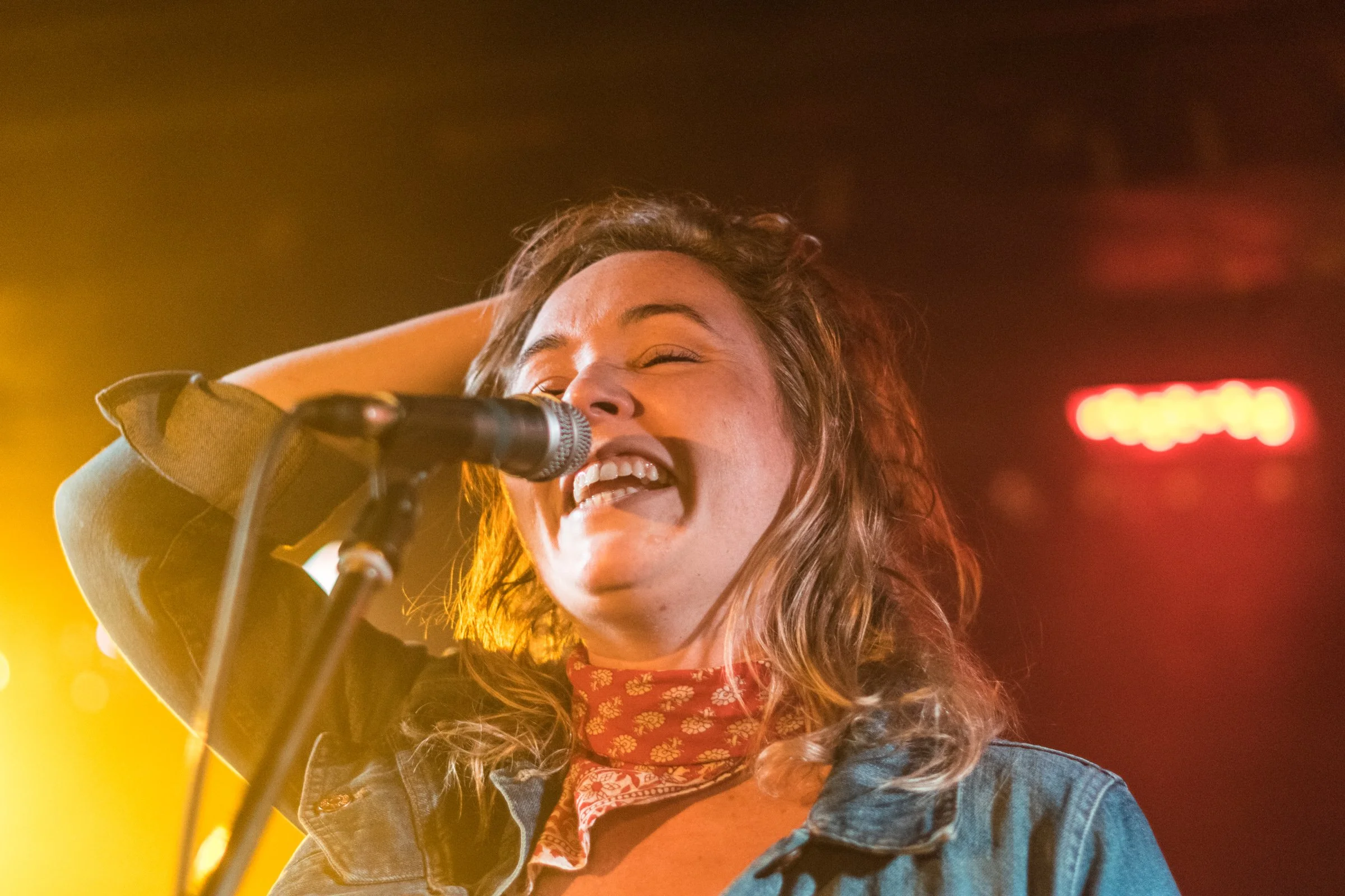A woman with curly hair singing into a microphone, smiling, with colorful lighting in the background.