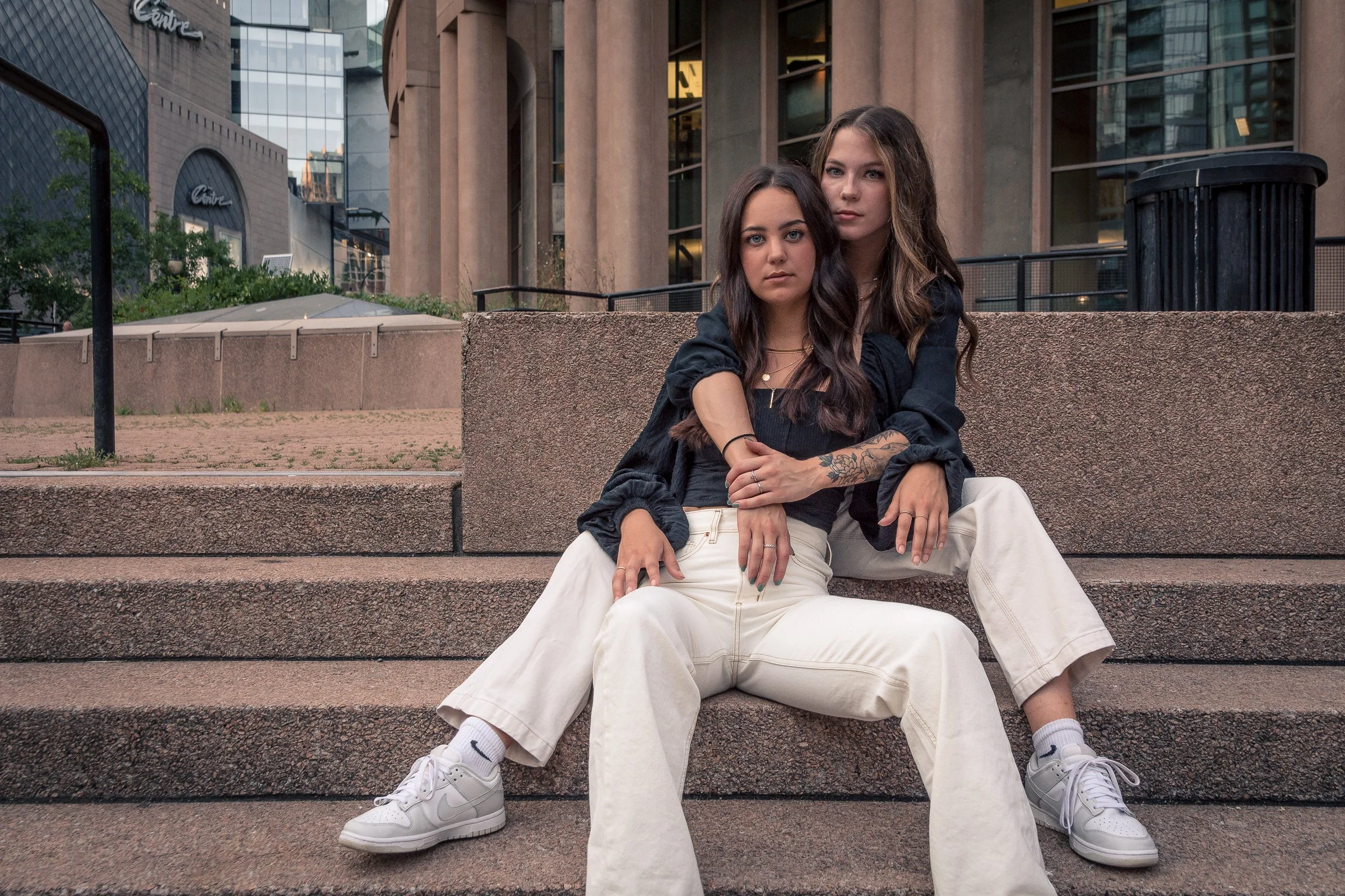 Two young women sit on concrete steps in an urban setting, with modern buildings in the background. They have long dark hair and are dressed casually, sitting close together, with one resting her arm on the other's shoulder.