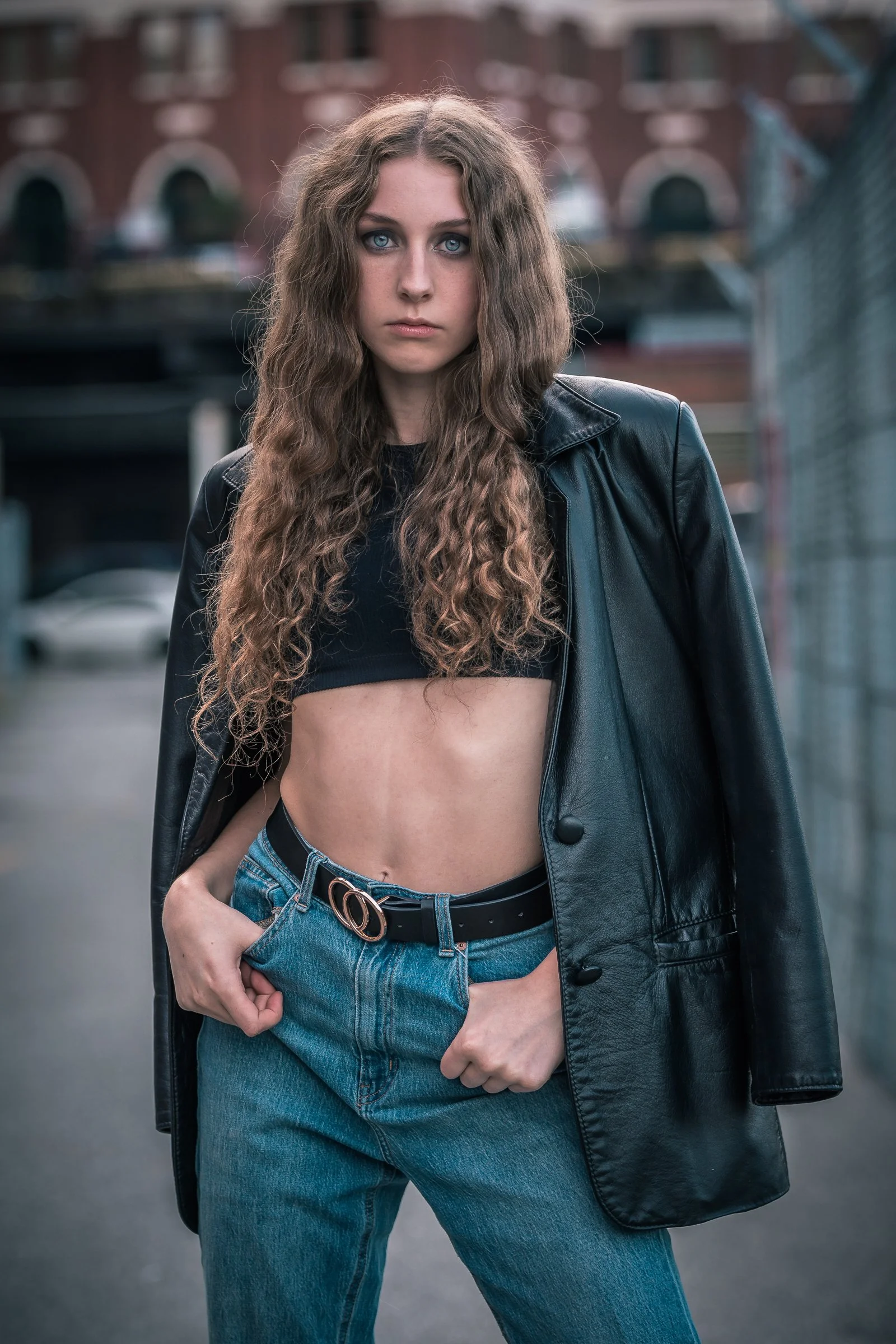 Young woman with long, curly hair and piercing blue eyes, wearing a black crop top, blue jeans with a belt, and a black leather jacket draped over her shoulders, standing outdoors in front of a blurred urban background.