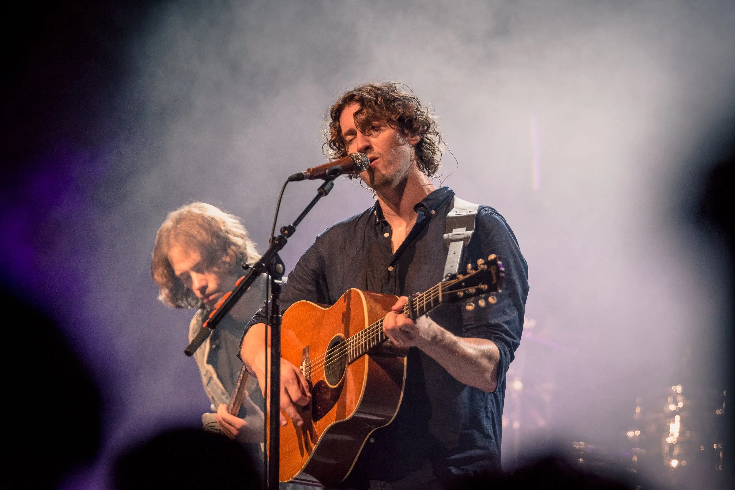A man with curly hair singing into a microphone while playing an acoustic guitar on stage in a performance setting, with another musician in the background.