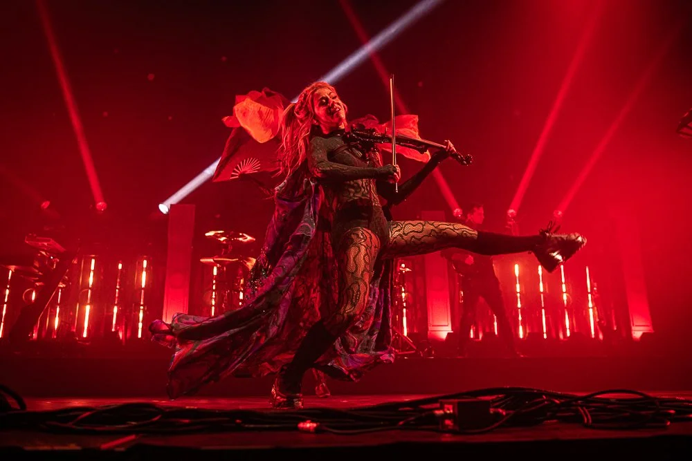 Performer in black lace outfit playing violin with props resembling wings, on stage illuminated by red lights, with backup musicians in the background.