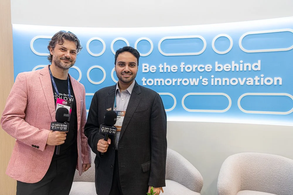 Two men standing with microphones in front of a bright blue backdrop that reads 'Be the force behind tomorrow's innovation.'