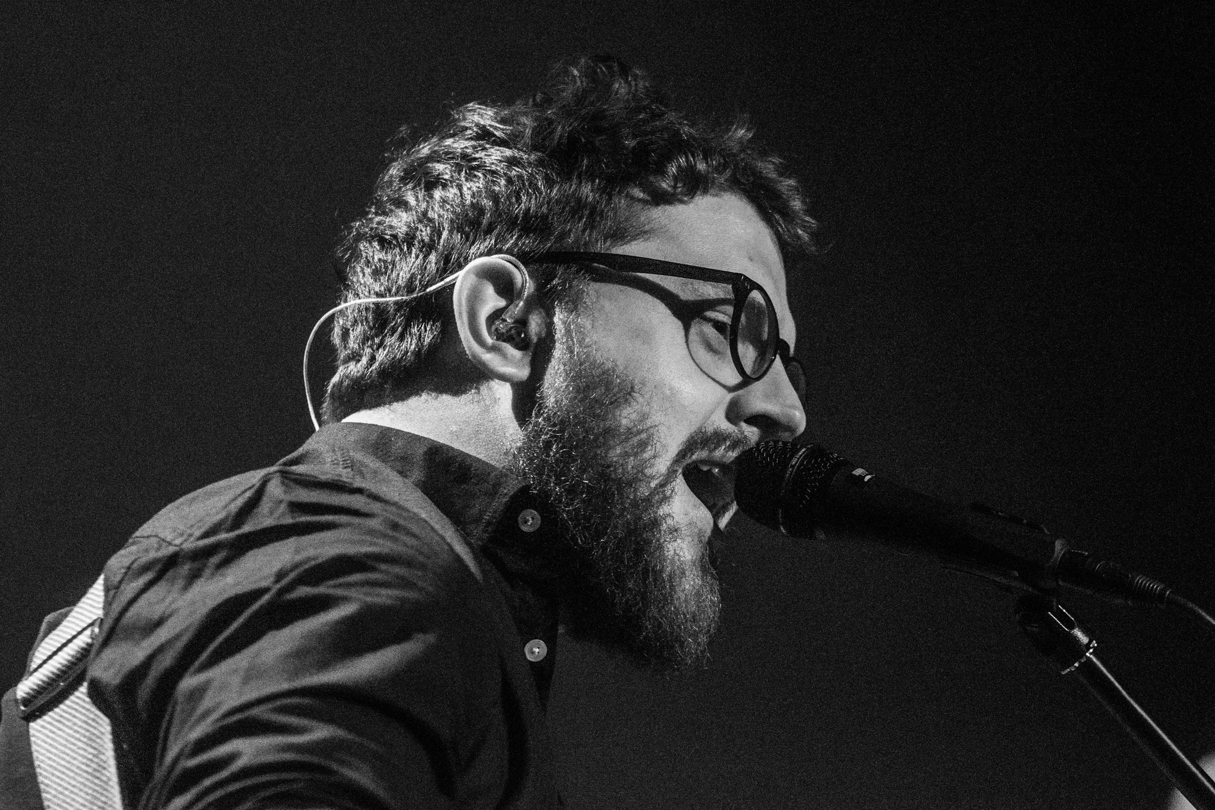 A black-and-white photograph of a male musician singing into a microphone on stage, wearing glasses and a jacket.