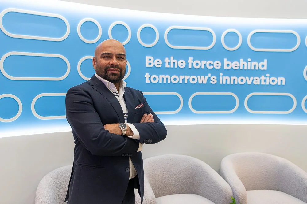 A man in a dark suit with a white shirt and watch standing with arms crossed in front of a blue wall with an inspiring quote.