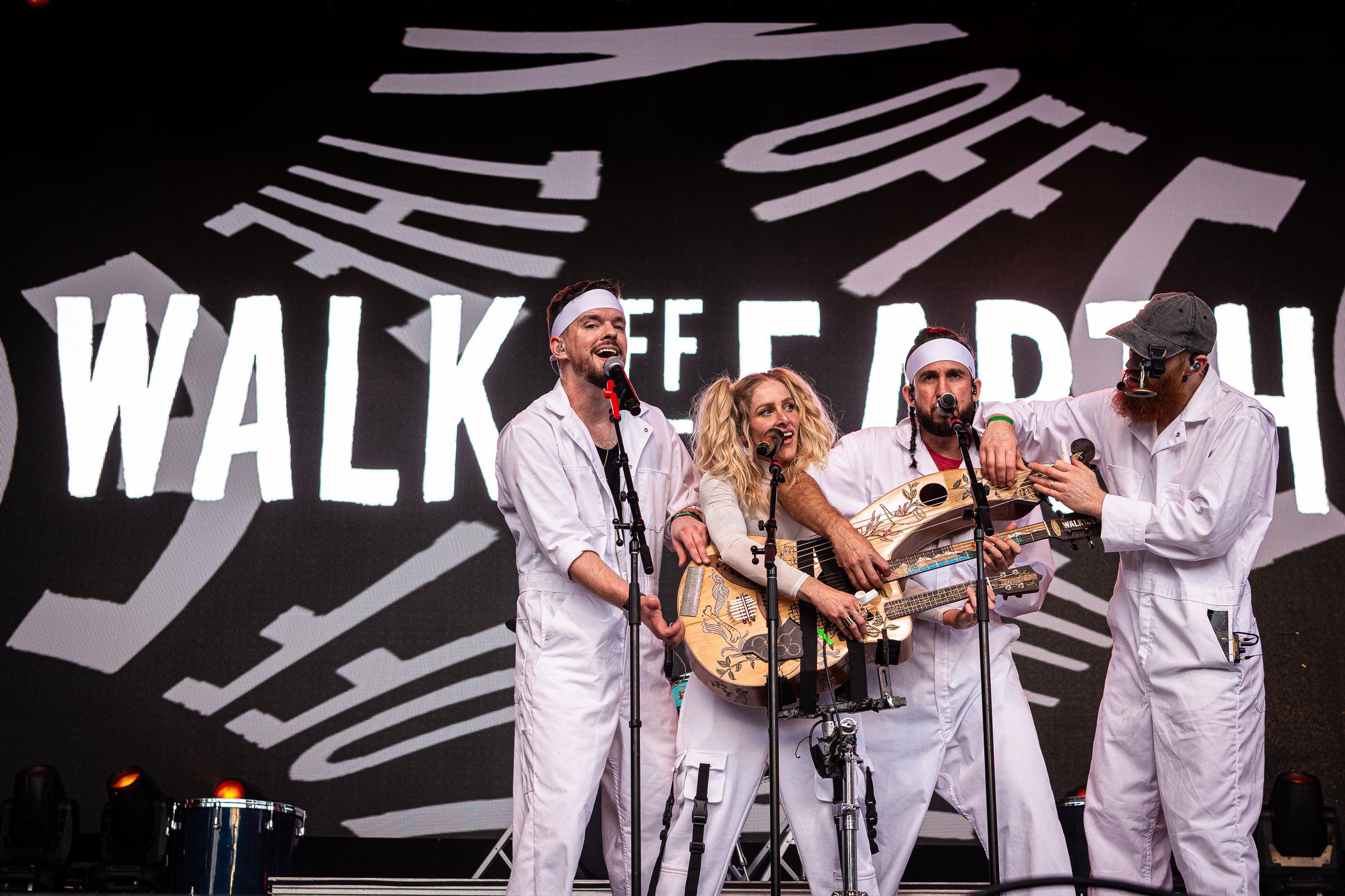 A group of four musicians dressed in white costumes performing on stage. One is holding a guitar, another appears to be singing, and the others are also engaged in singing or playing instruments. Behind them, a large black and white graphic with the 