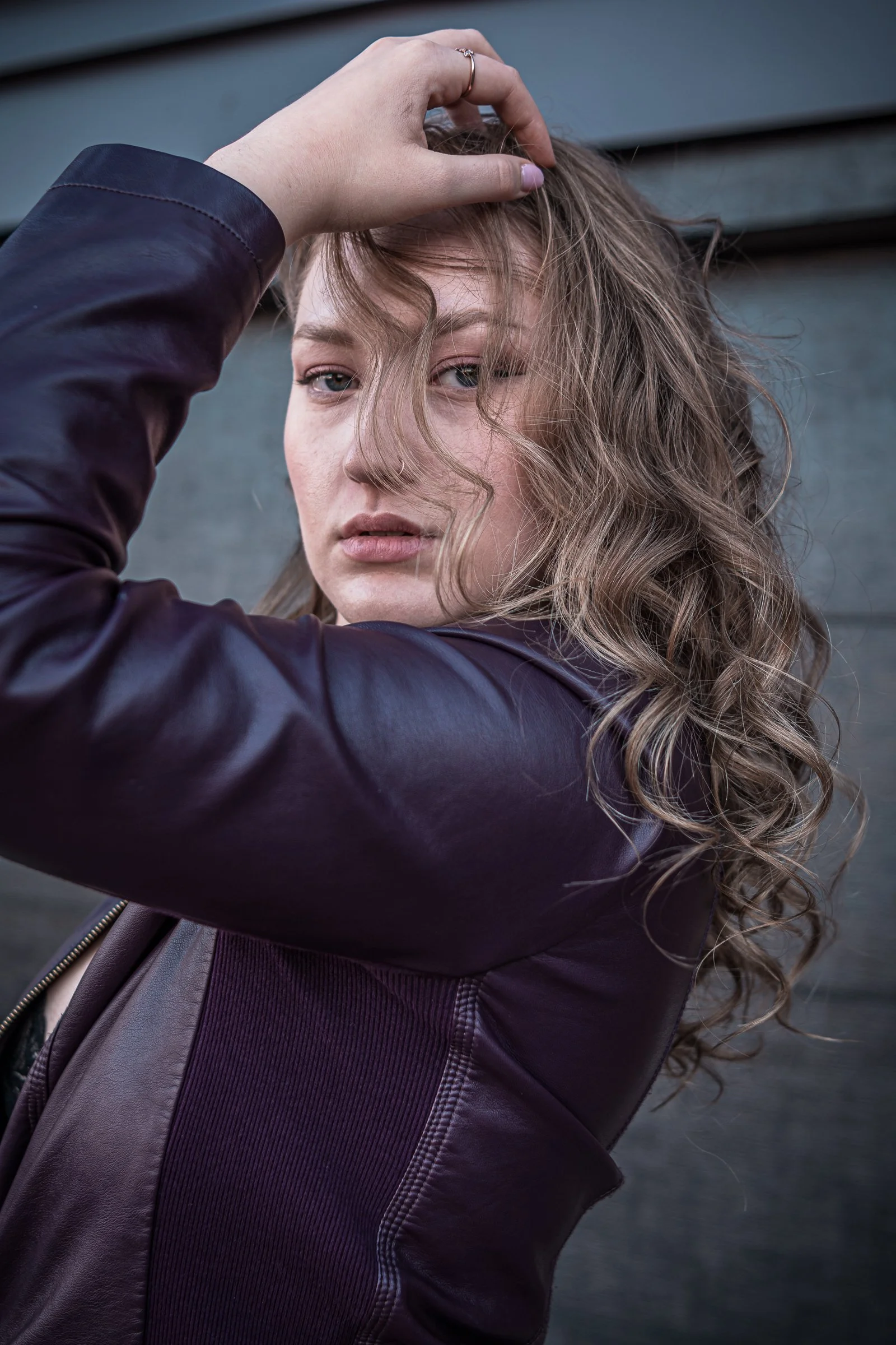 Close-up of a woman with curly hair, wearing a leather jacket, touching her hair with one hand, in front of a grey wall.