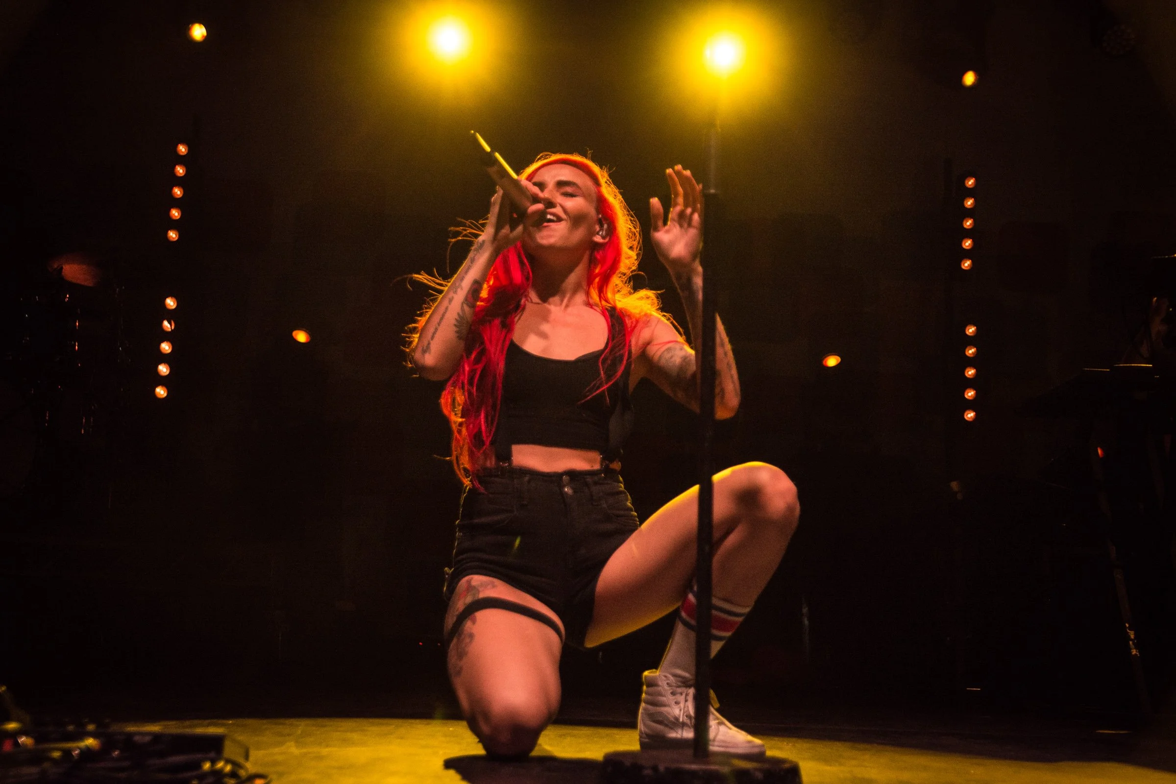 A woman with red hair singing into a microphone on stage, wearing a black crop top and shorts, with tattoos on her arms, illuminated by yellow stage lights.