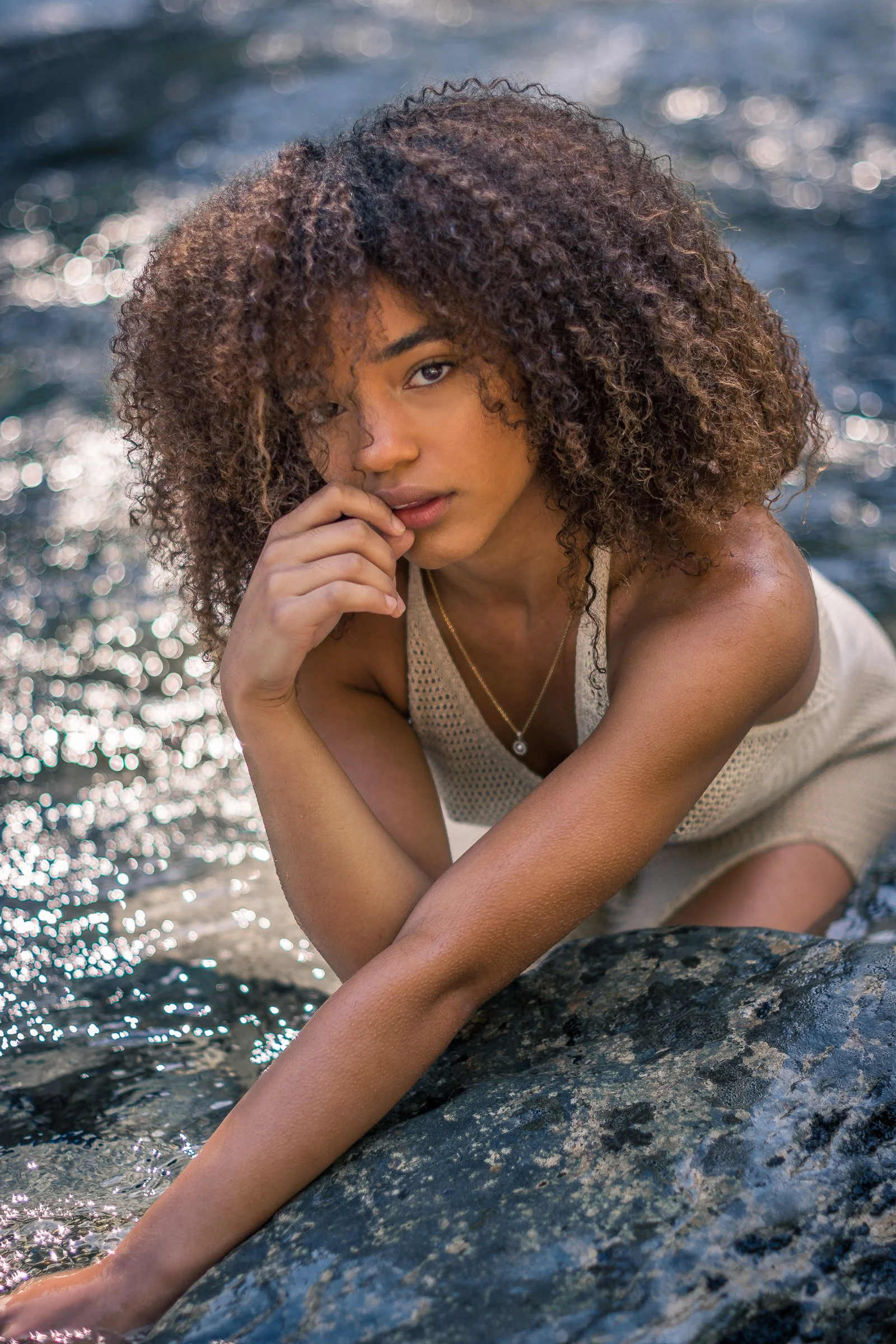 A young woman with curly hair faces the camera while sitting on rocks near water, with sunlight reflecting off the water.