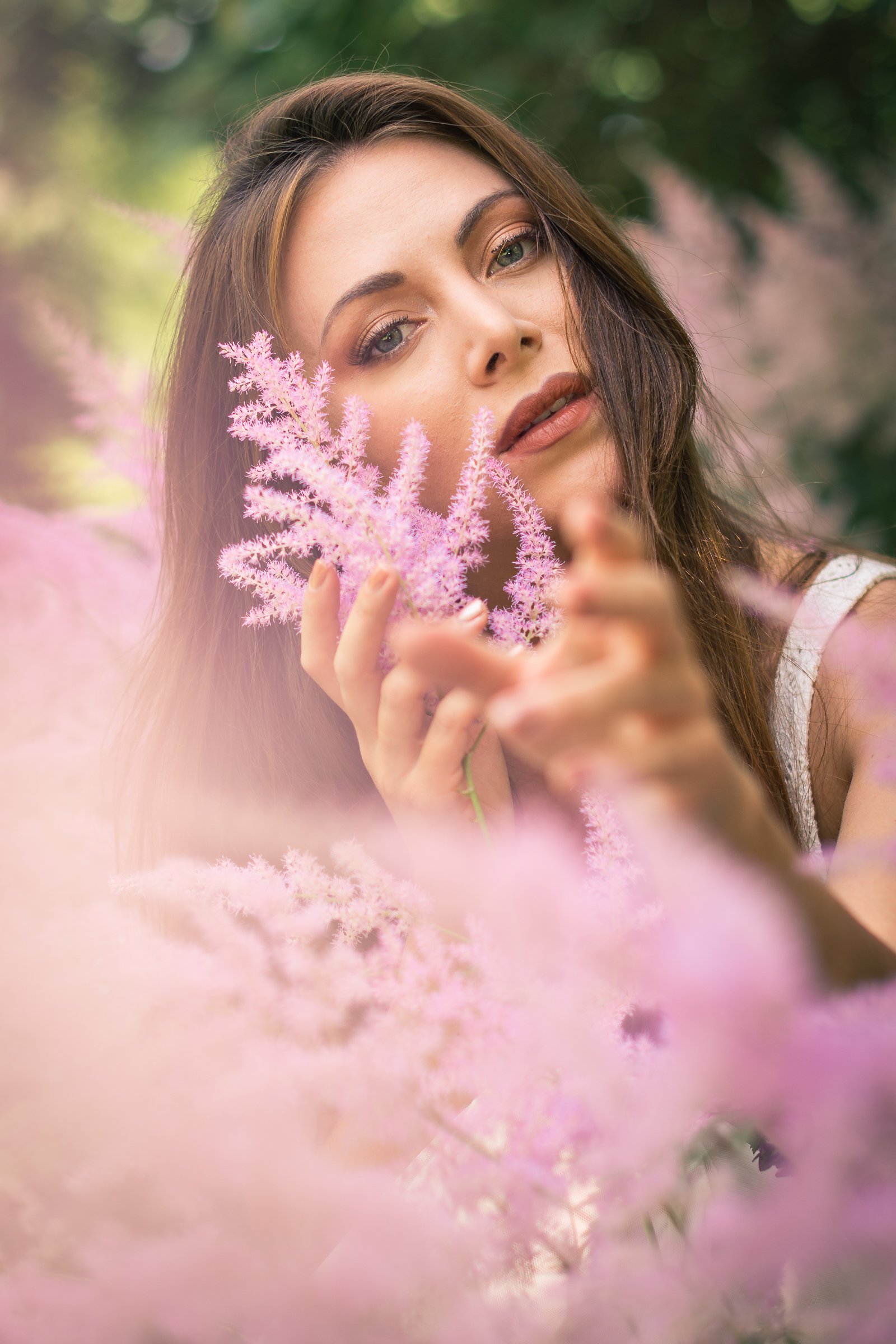 A woman with brown hair and green eyes holding a pink fluffy flower close to her face outdoors.