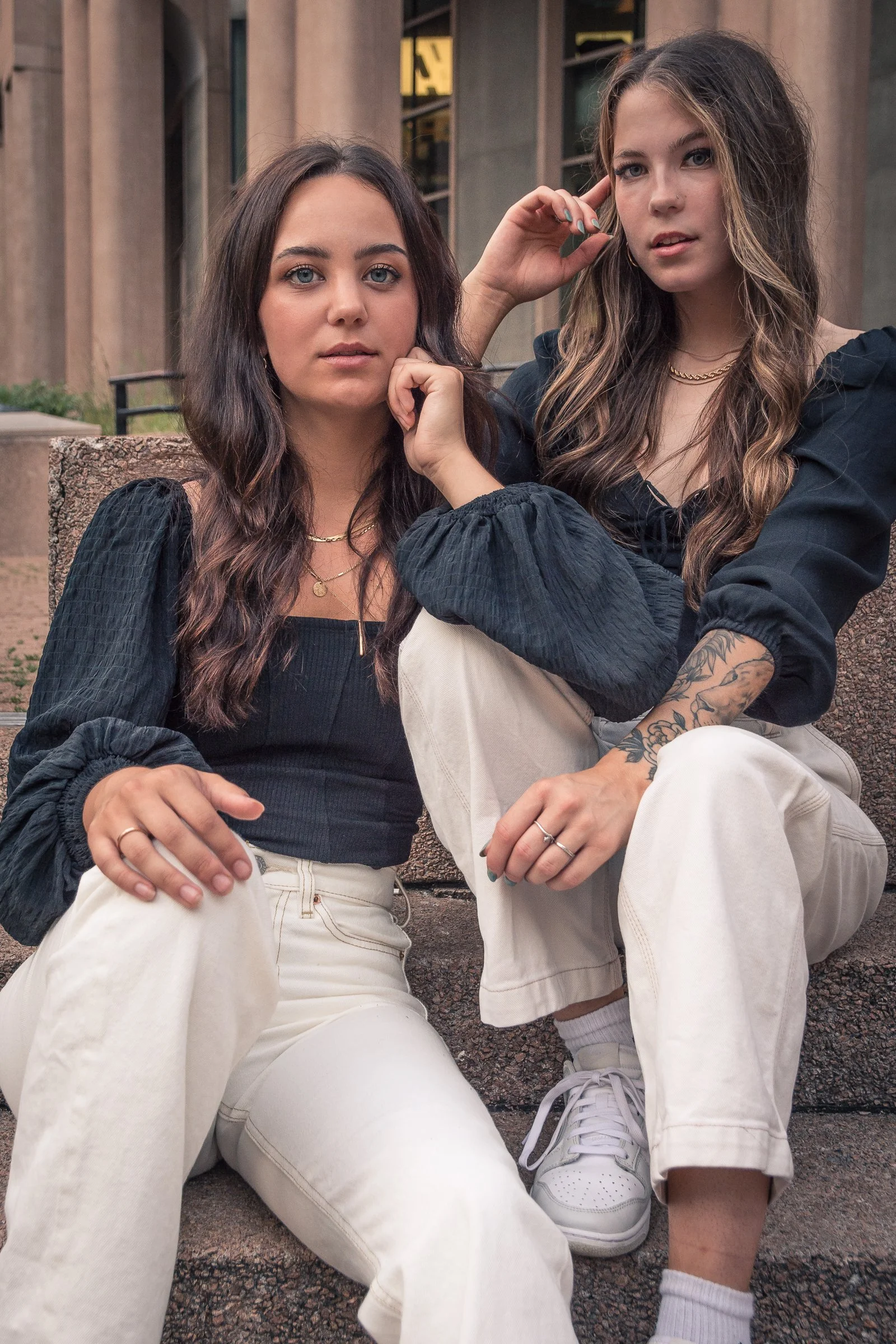 Two young women sitting outdoors on a stone bench, facing the camera, with modern building architecture in the background. Both are dressed casually in light-colored pants and dark tops, with one woman wearing white sneakers and the other in white so