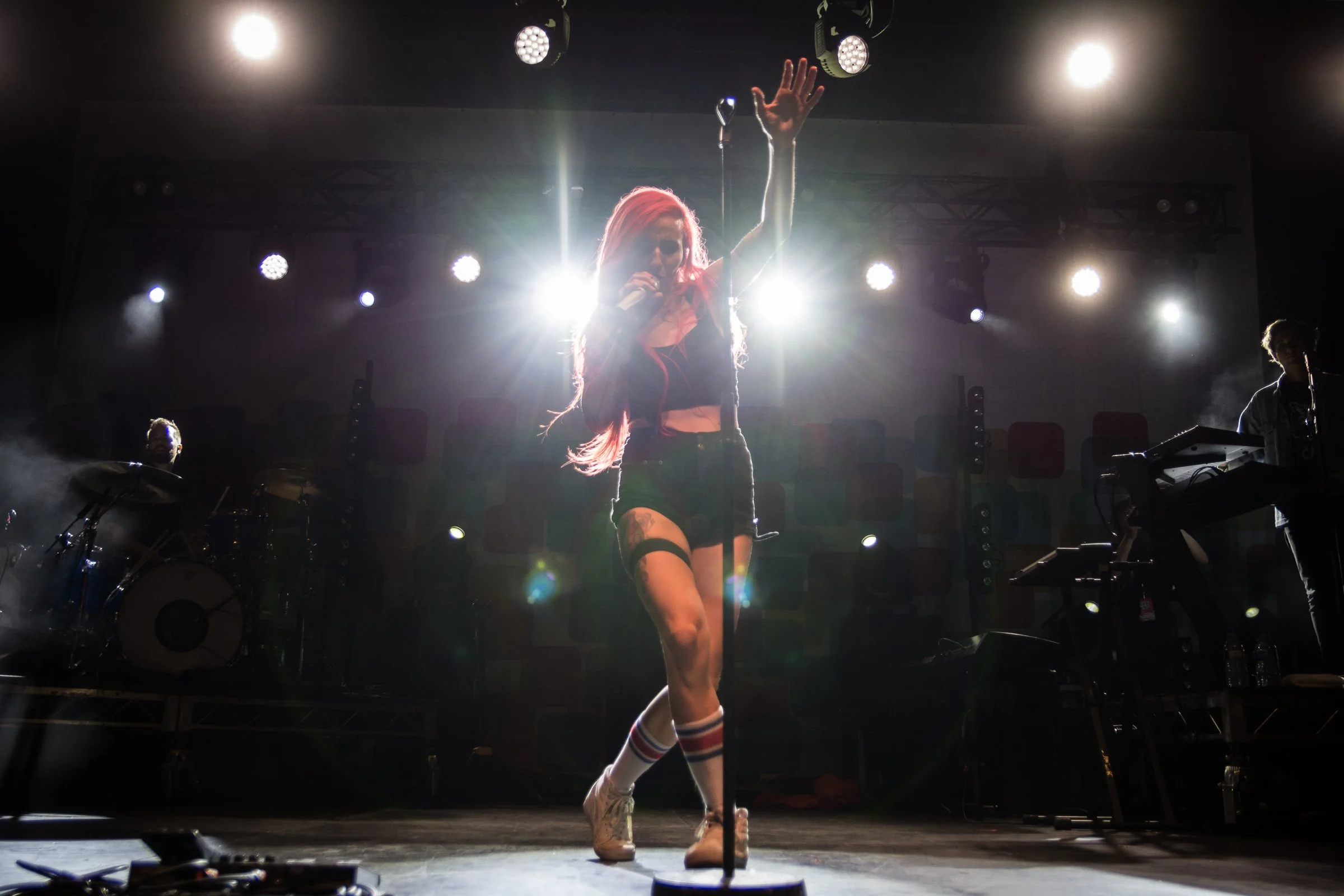 A female singer with pink hair singing into a microphone on stage, with bright stage lights and musicians in the background.