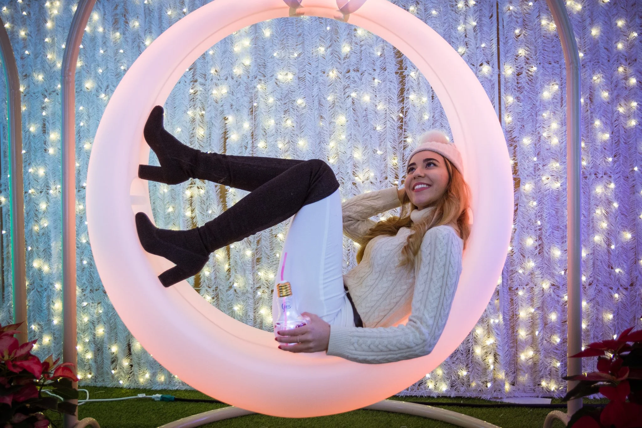 A young woman with long hair and a knit beanie hat smiling while lying on an illuminated circular light with a snowy, Christmas-themed background featuring white pine branches with fairy lights.