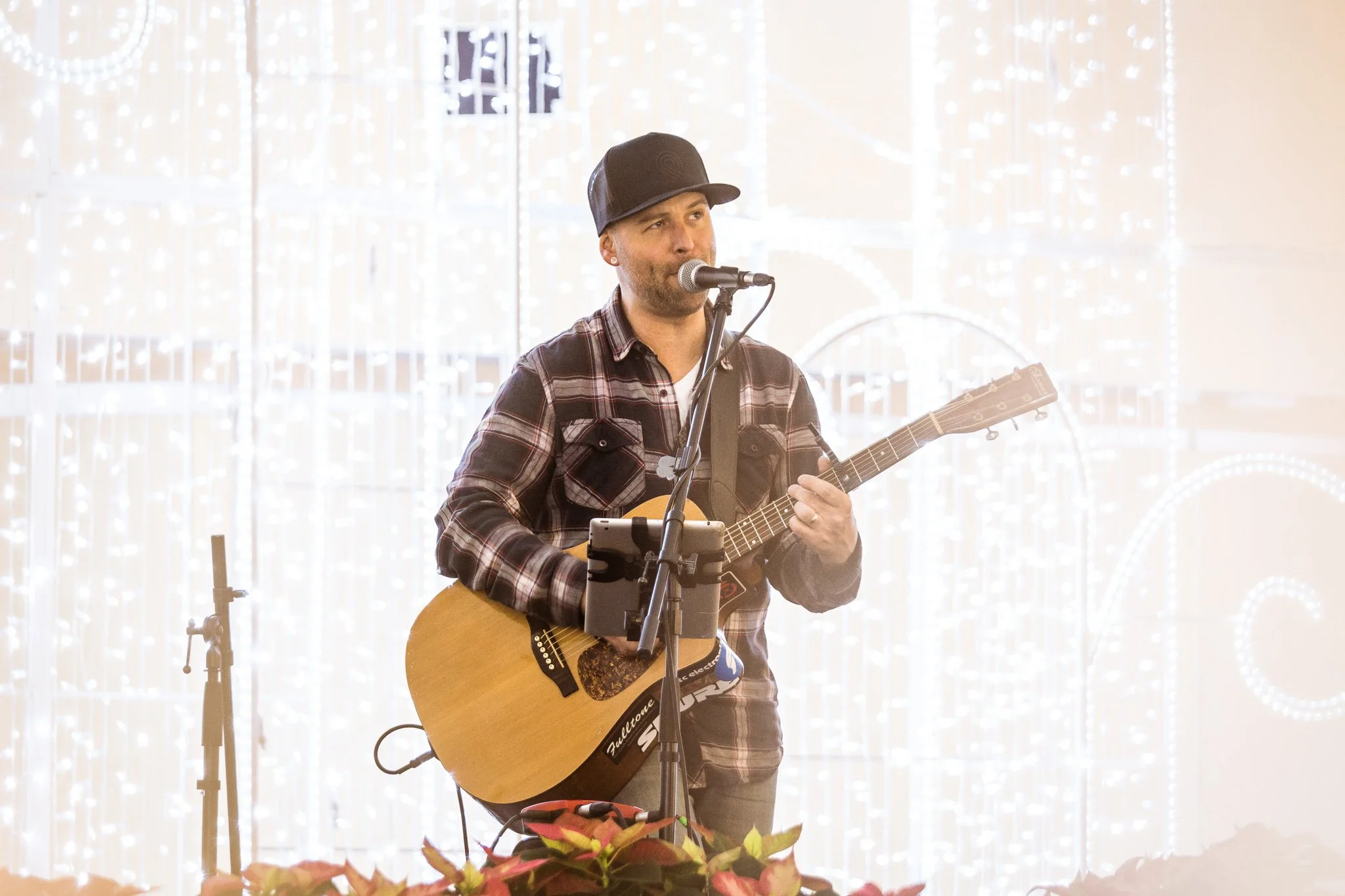 A man wearing a black baseball cap and a plaid shirt singing and playing an acoustic guitar at a performance, with Christmas lights in the background.