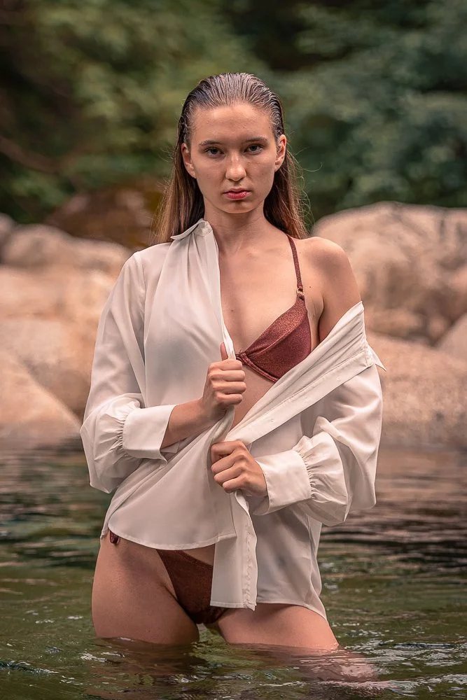 A young woman standing in a body of water, holding open a white shirt to reveal a brown bra and matching underwear, with a rocky and wooded background.