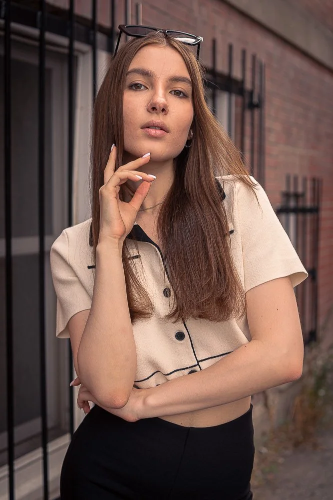 Young woman with long brown hair wearing glasses on her head, a beige blouse, and black pants, standing outdoors near a brick building.