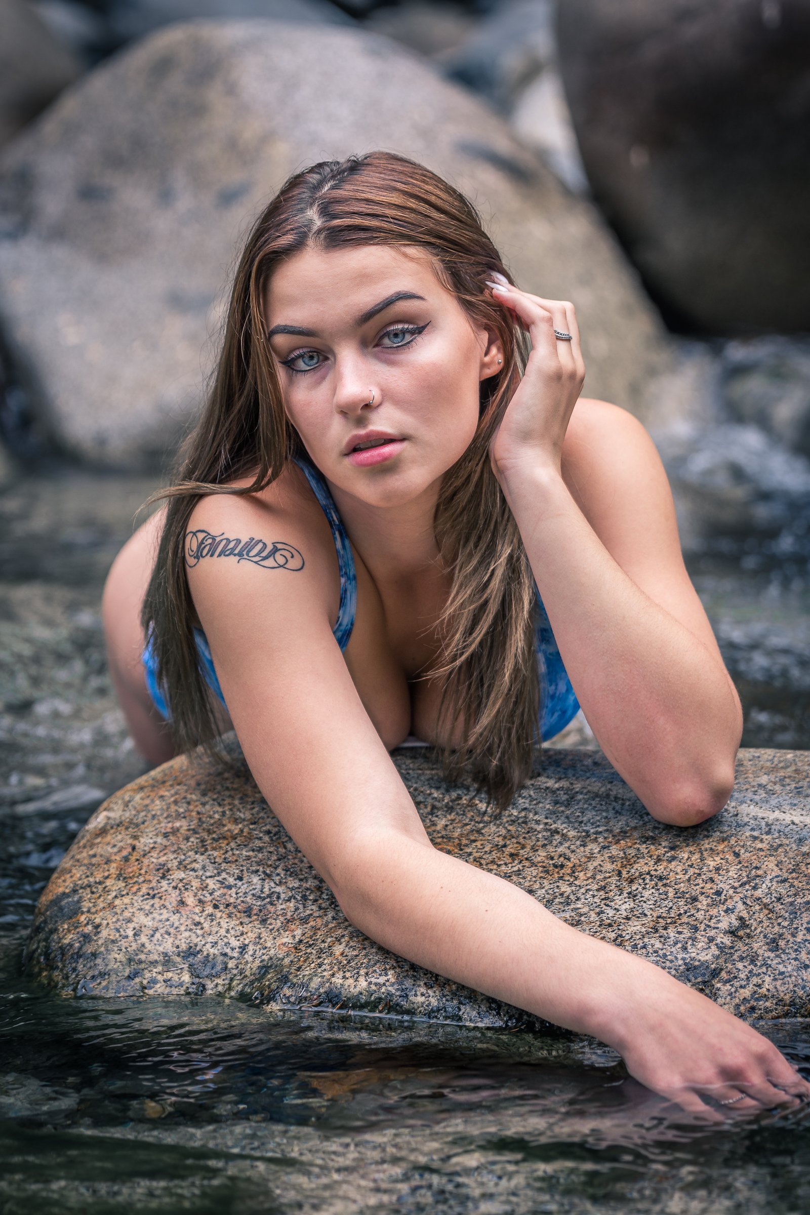 A young woman with blue eyes has long brown hair and is lying on a large rock near a body of water, with more rocks in the background. She has a tattoo on her shoulder and is touching her ear with one hand.