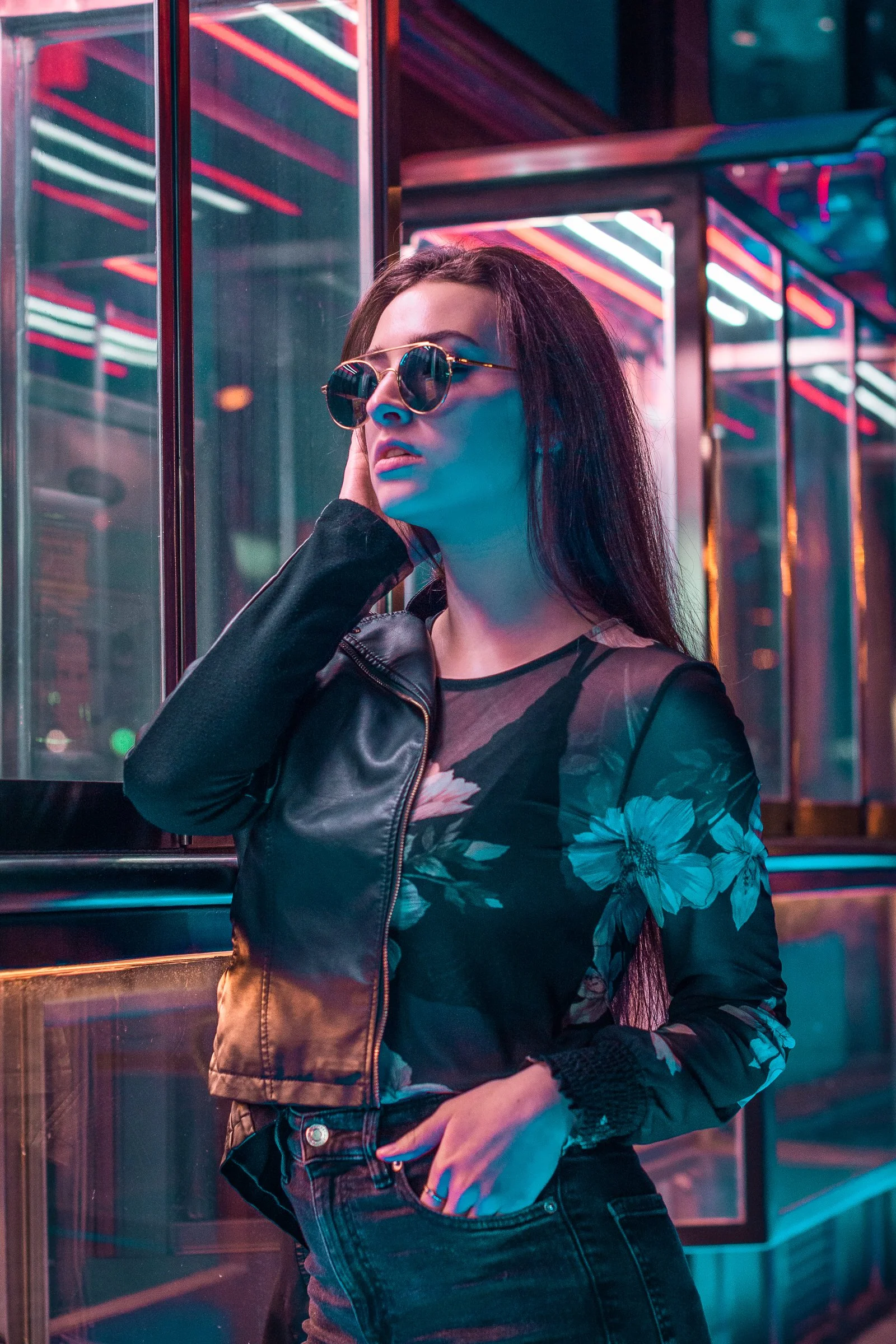 A woman wearing round sunglasses, a floral-printed top, and a black leather jacket, standing in front of a glass wall illuminated by colorful neon lights, with her hand touching her face.