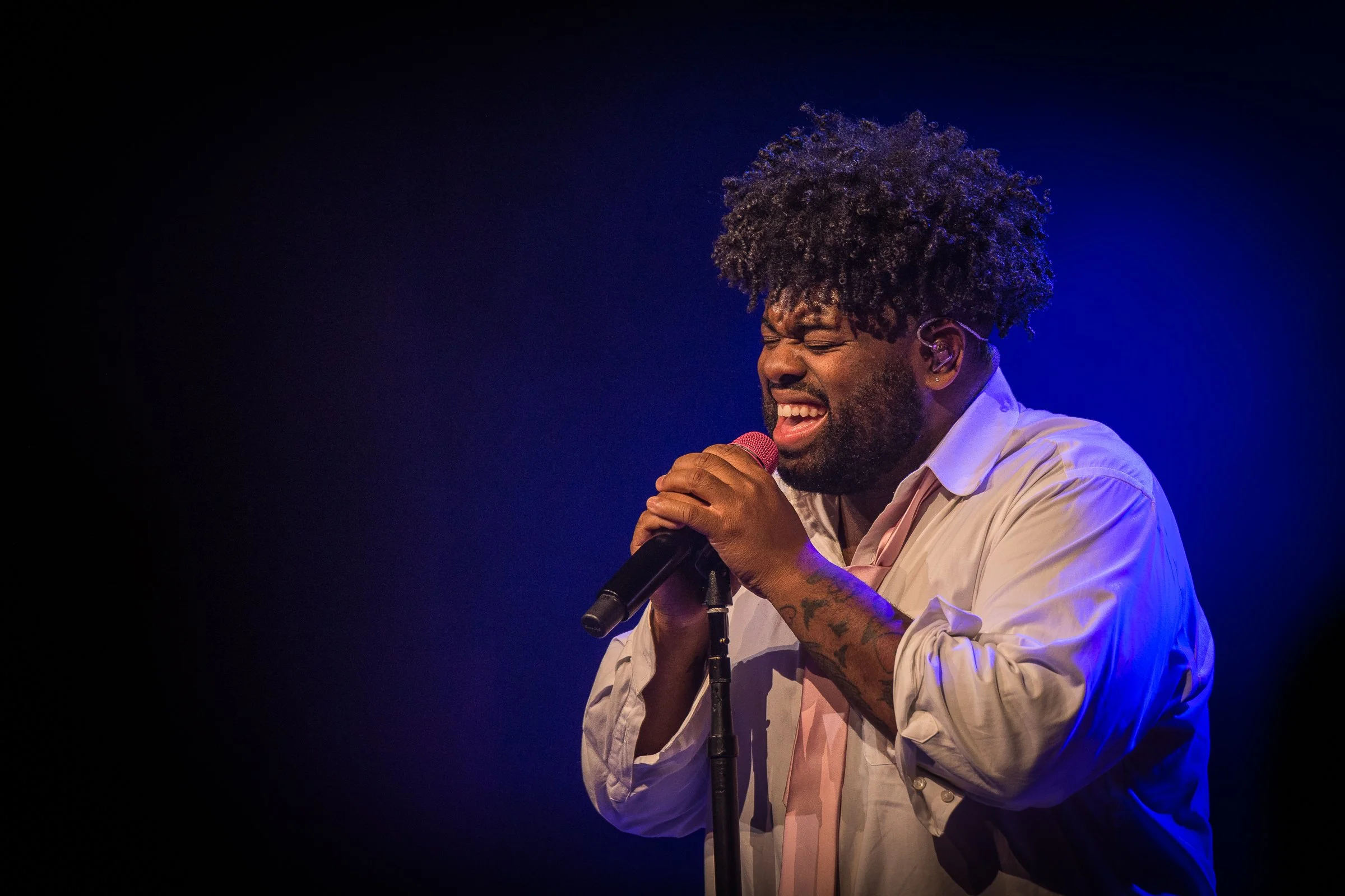 A man singing passionately into a microphone, eyes closed, wearing a white shirt with pink tie, standing on stage with a dark background and blue and purple stage lighting.