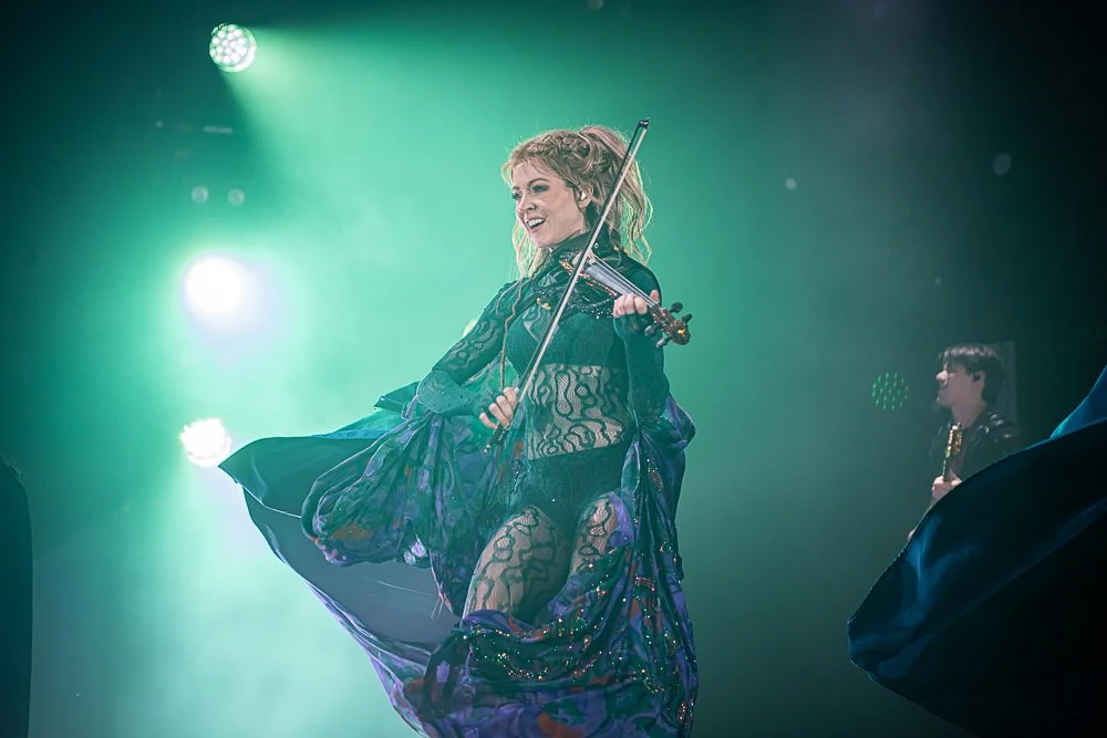 A woman in a black lace outfit plays a violin on stage, smiling with a flowing purple and green skirt, with green stage lights in the background.