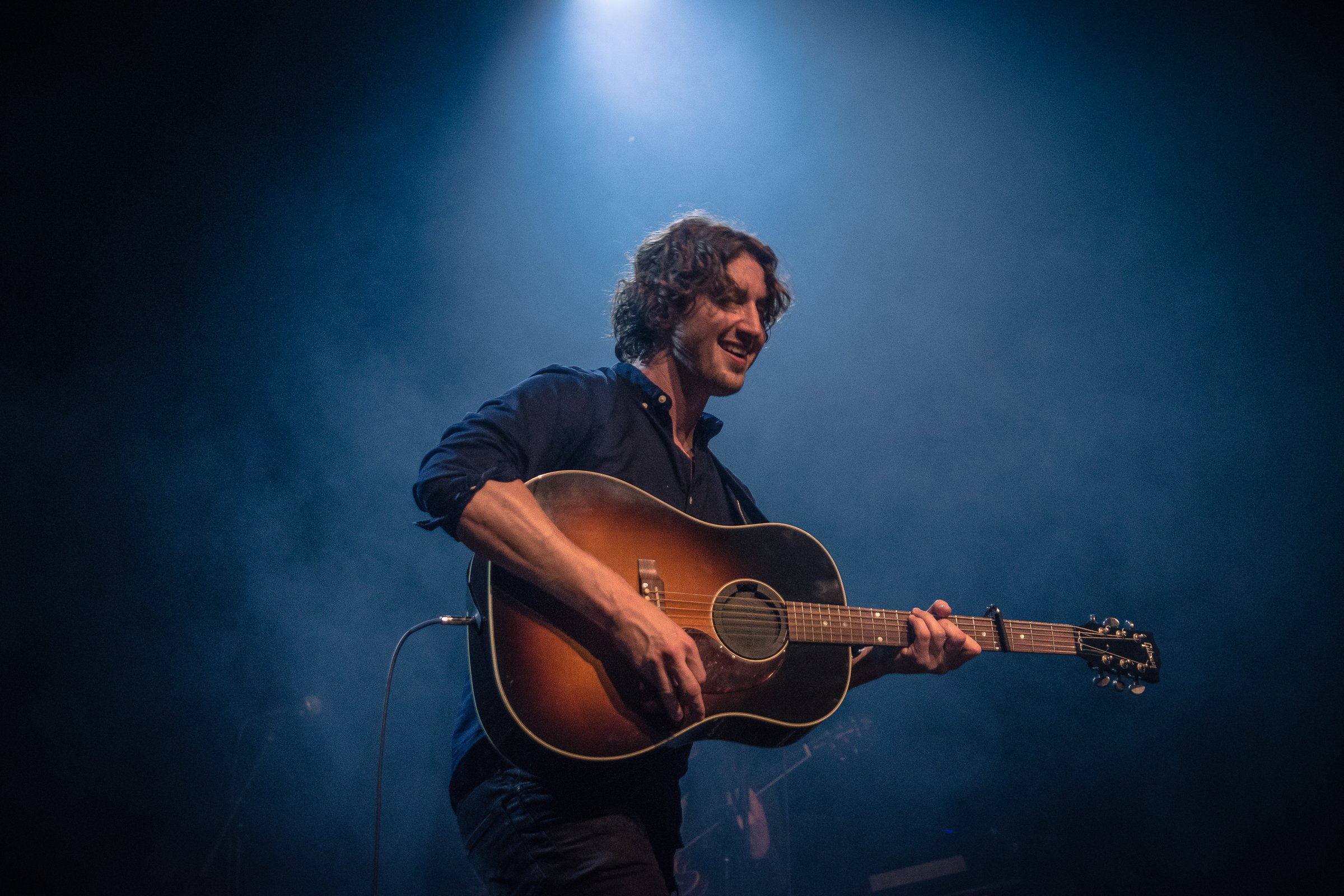 A man with curly dark hair smiling and playing an acoustic guitar on stage under blue lighting.