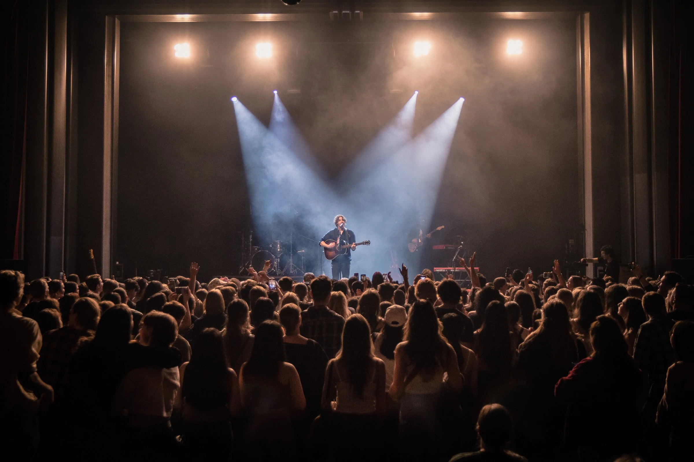 A male musician with curly hair playing an acoustic guitar on stage, performing in front of a large audience at a concert. The stage is lit with spotlights, and the audience has their hands raised.