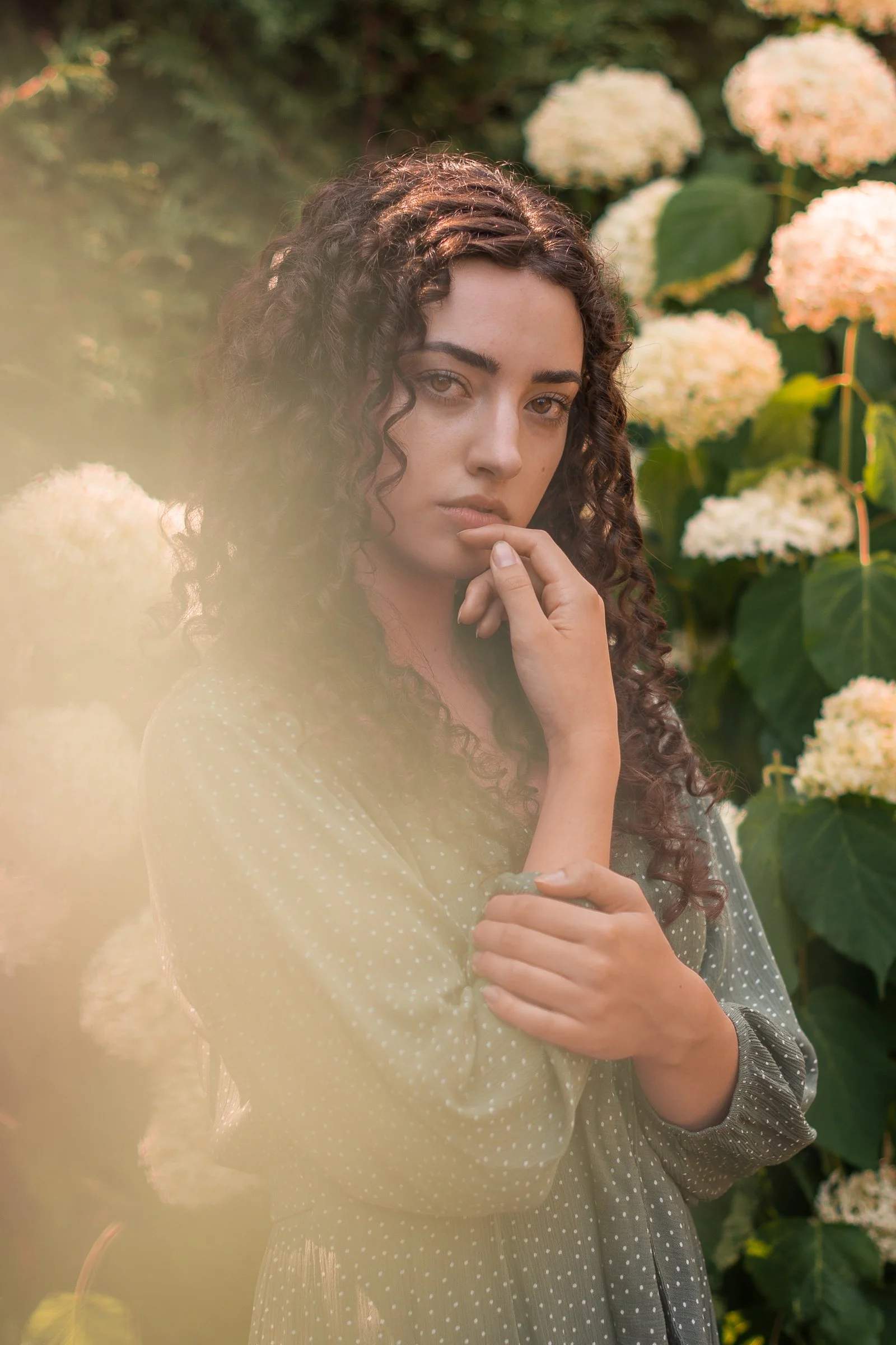 A woman with curly hair standing amongst bushy plants with white flowers, wearing a light-colored, polka-dot dress and a sheer scarf.