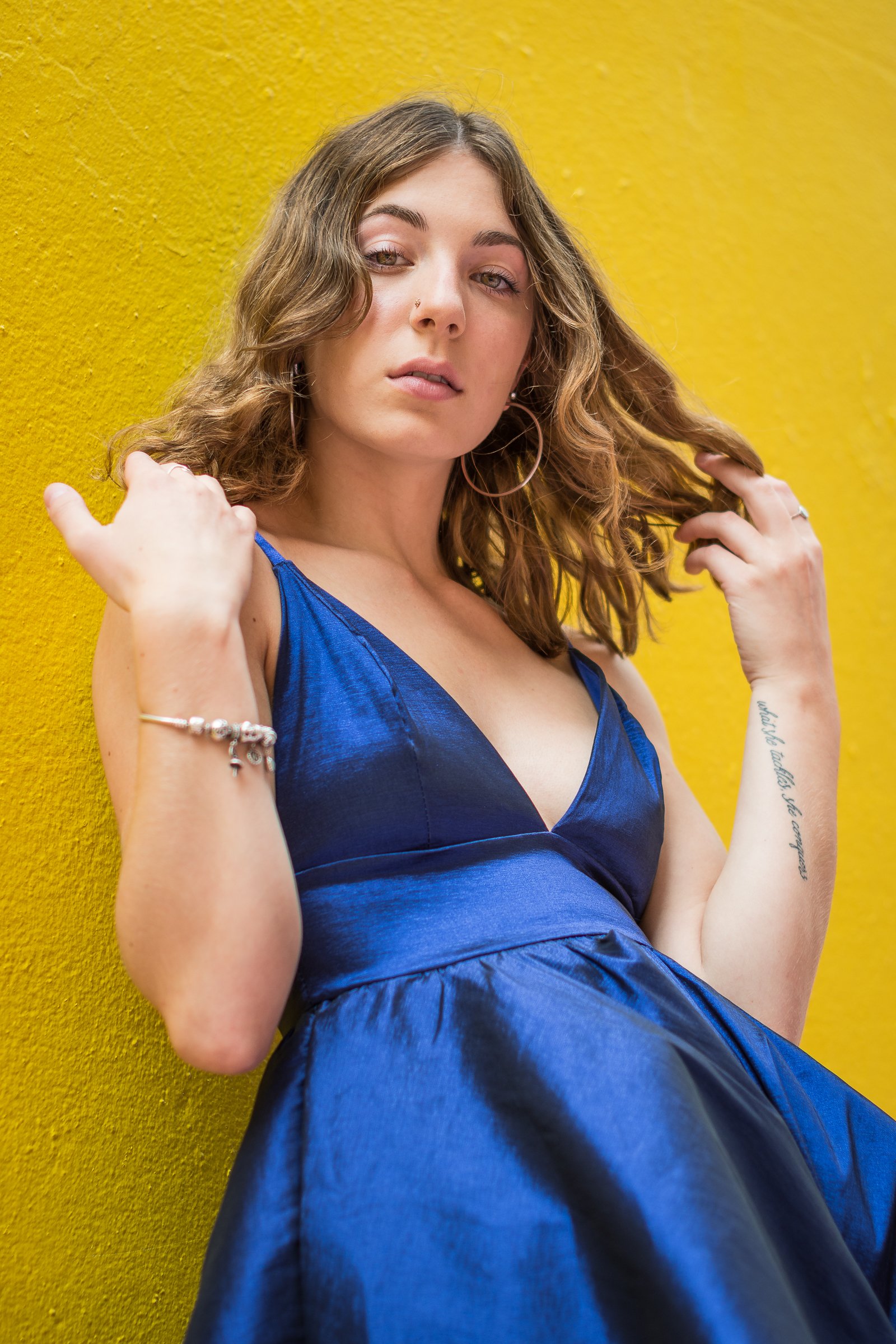 A young woman with wavy brown hair and hoop earrings poses against a yellow wall, wearing a blue dress and jewelry.
