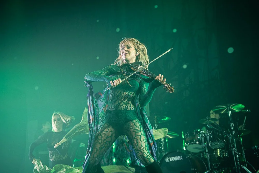 A woman playing an electric violin on stage, dressed in black lace clothing, with backup musicians and drums visible in the background, performing under green stage lighting.
