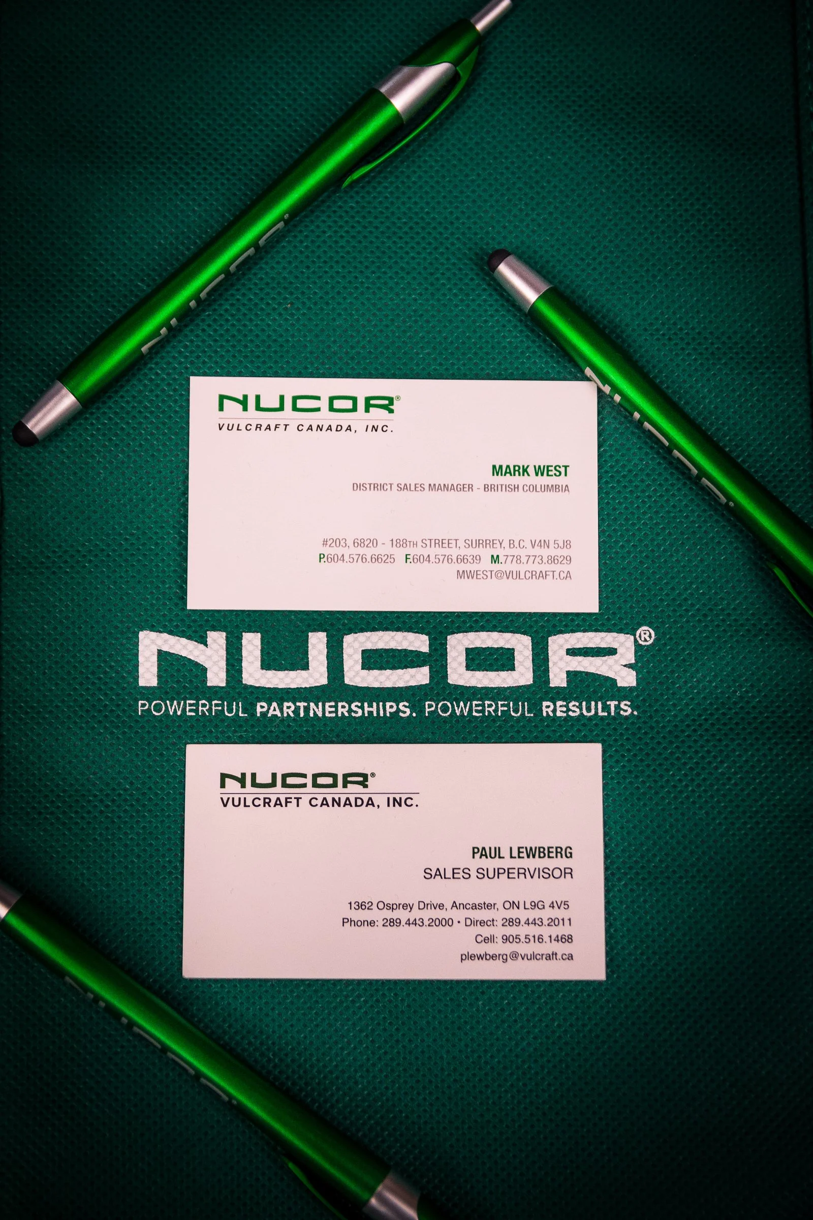Business cards and pens placed on a green textured background for NUCOR, Vulcraft Canada, Inc., featuring contact information and titles for Mark West and Paul Lewberg.