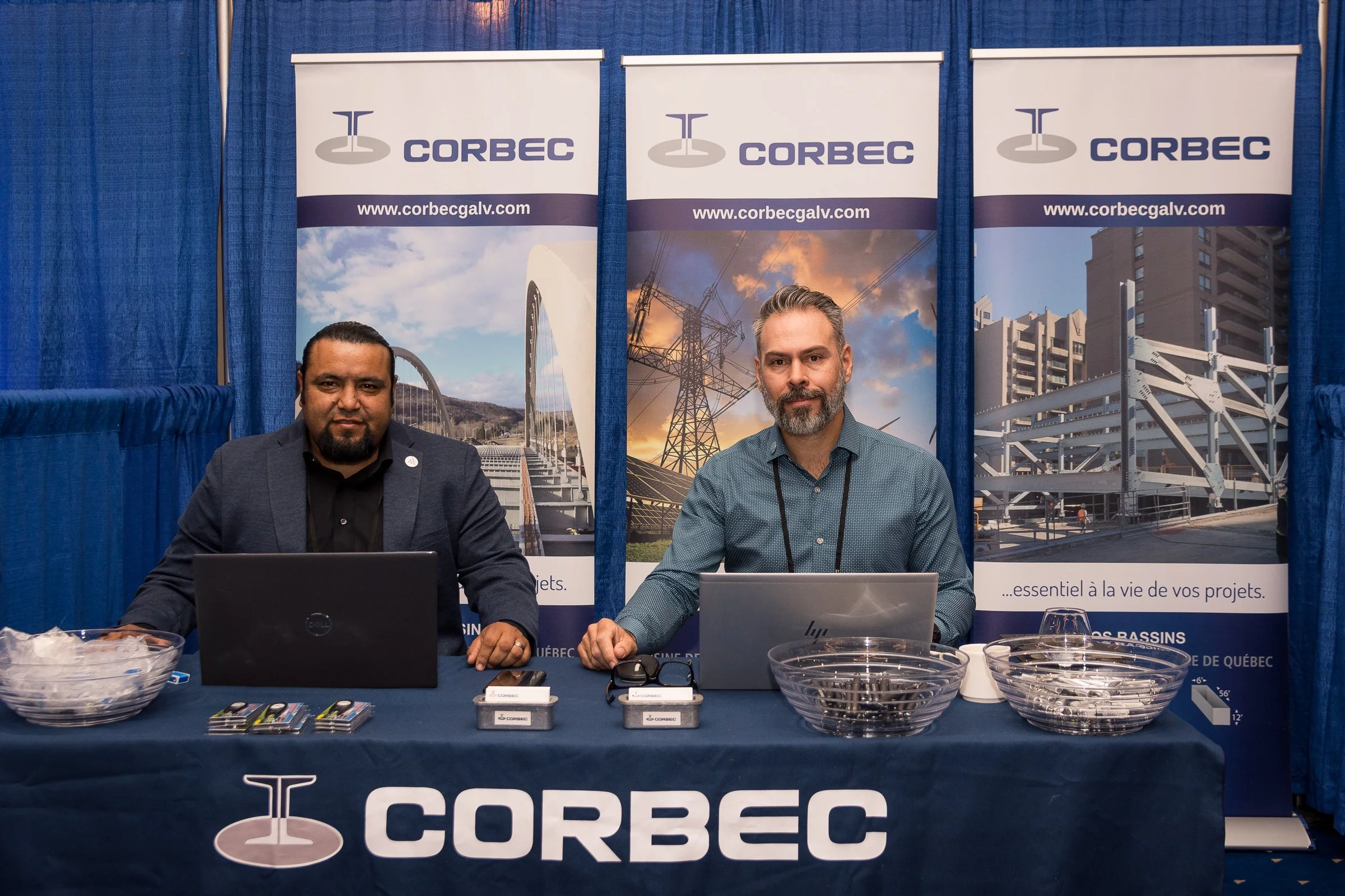 Two men sitting at a table with promotional banners behind them for CORBEC, a company specializing in electrical infrastructure. The table has laptops, business cards, and bowls of candies.
