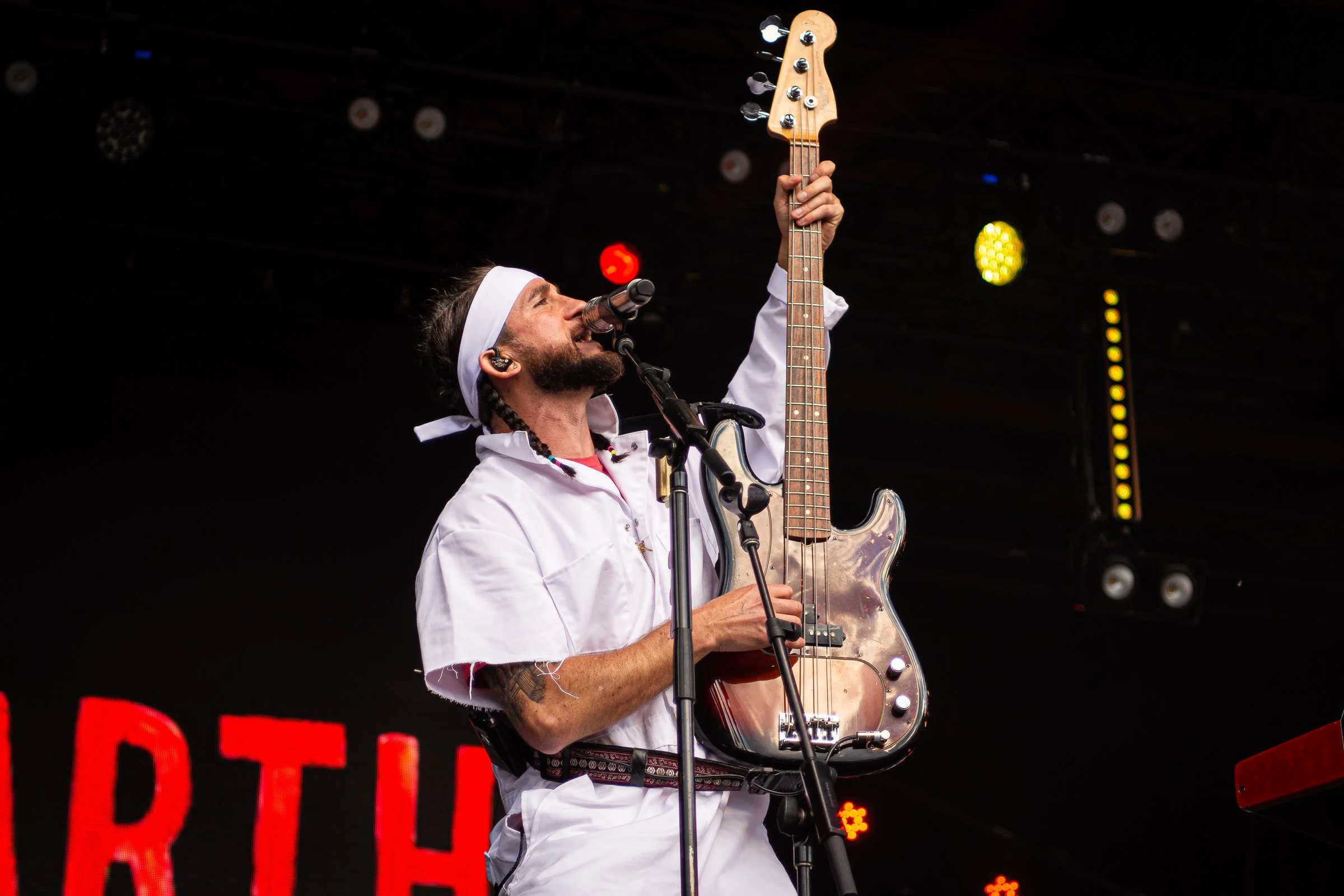 A man with braided hair wearing a white headband and shirt, playing an electric guitar on stage with a microphone in front of him.