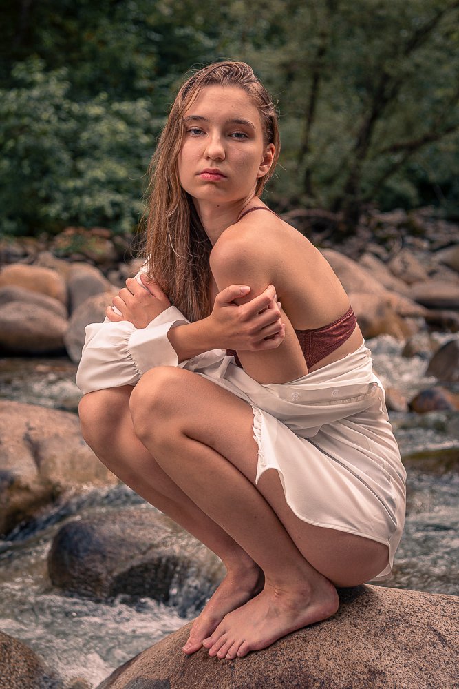 Young woman crouching on a rock by a stream in a forest, wearing a white dress and a brown top, with long hair and a serious expression.