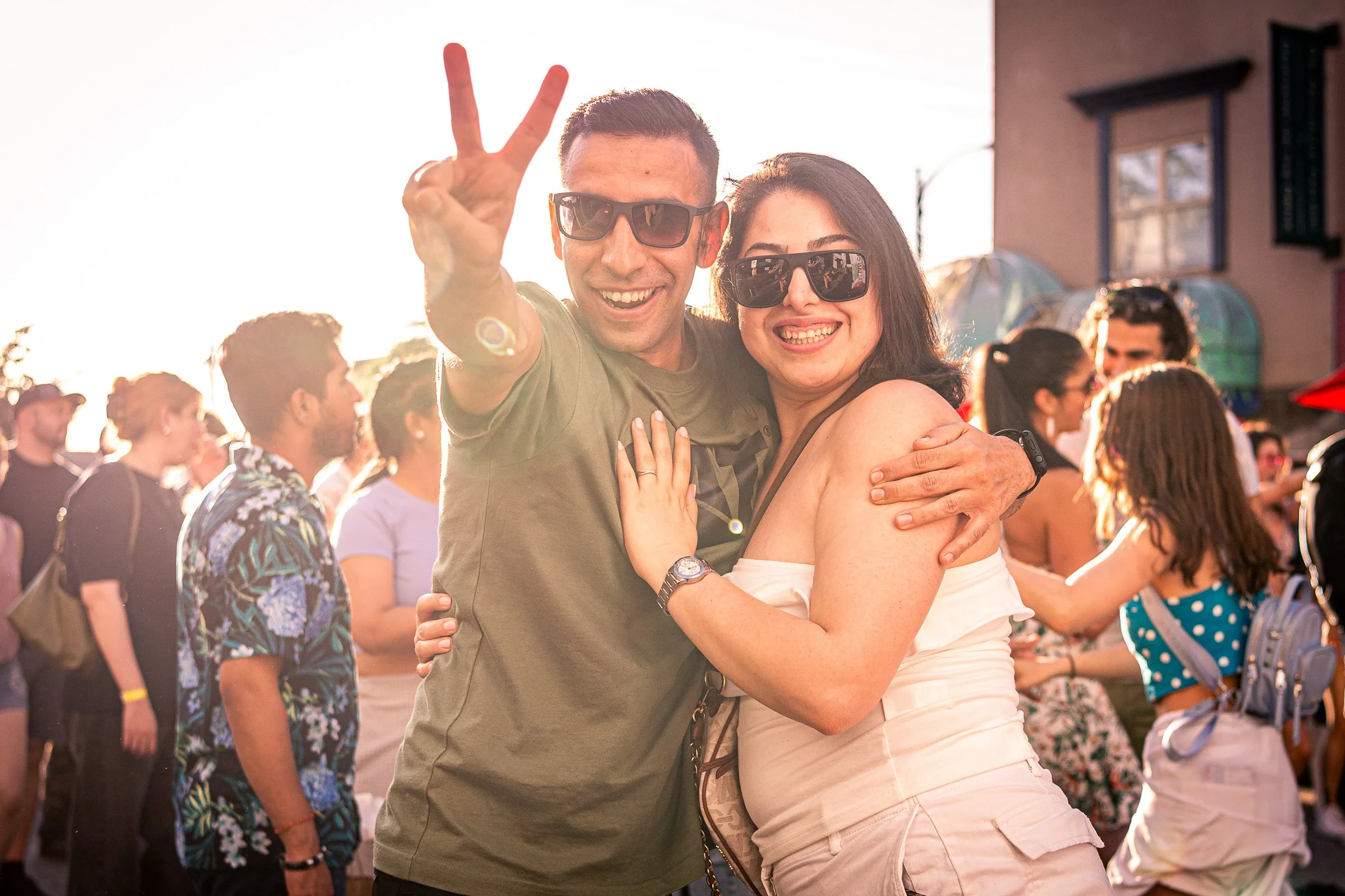 A smiling man and woman wearing sunglasses hugging at an outdoor event with other people in the background.
