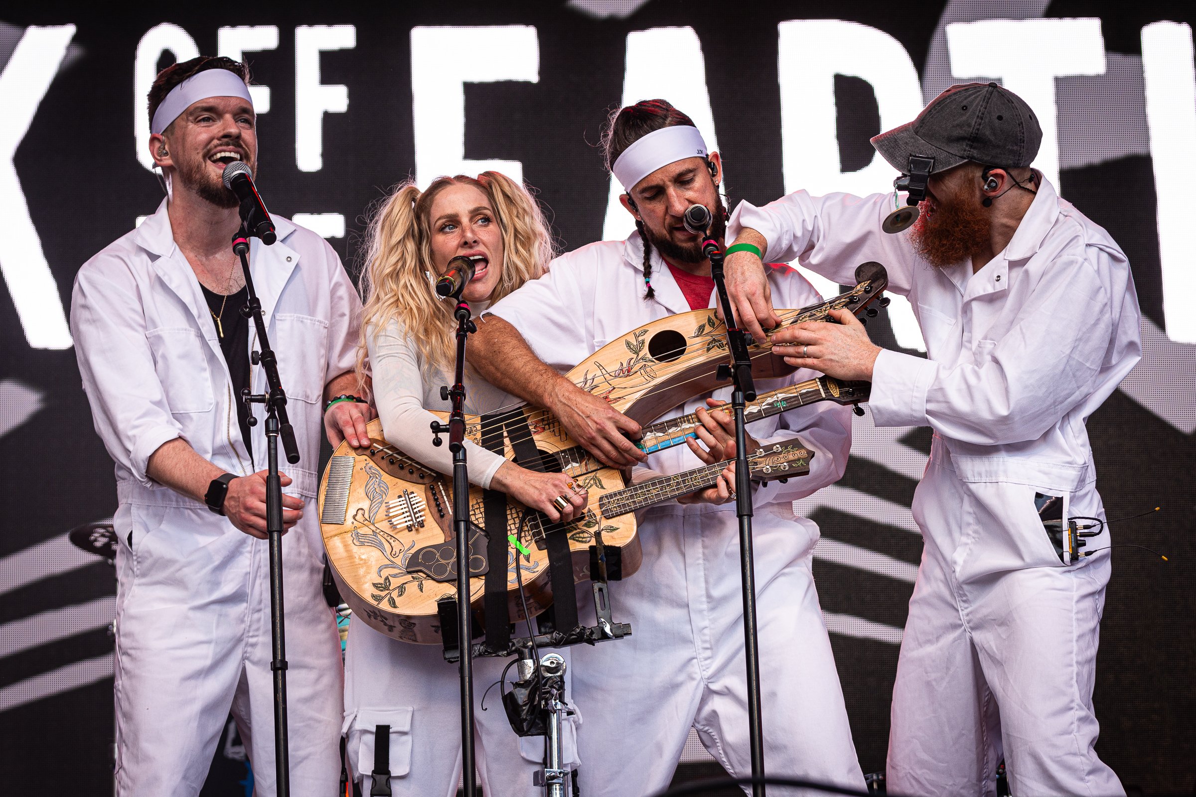 Four people dressed in white clothing performing on stage, with a large screen in the background displaying the text 'FREE.'