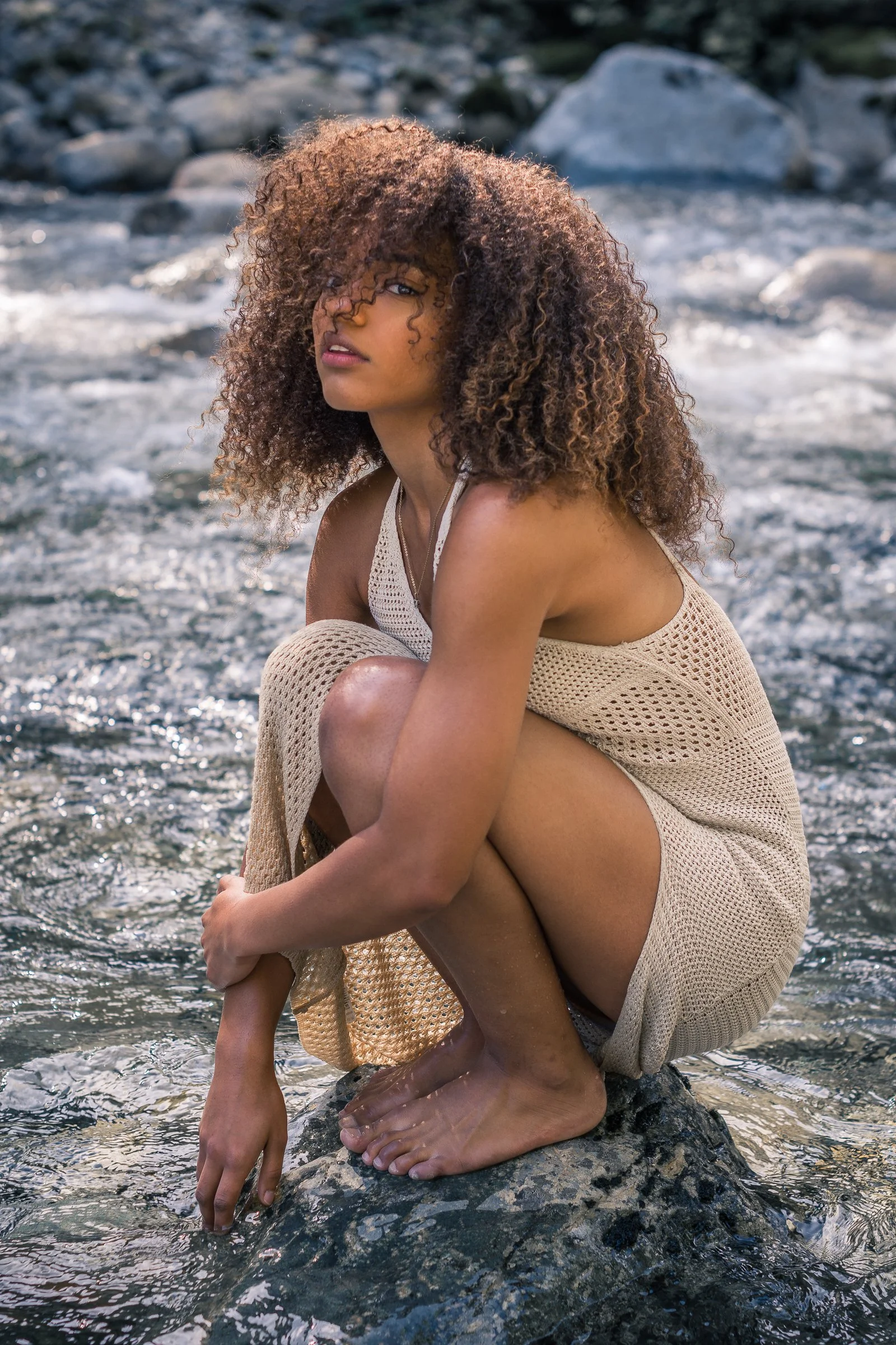 A woman with curly hair sitting on a rock in a shallow body of water, with one hand touching the water and the other arm wrapped around her knees, wearing a beige outfit.
