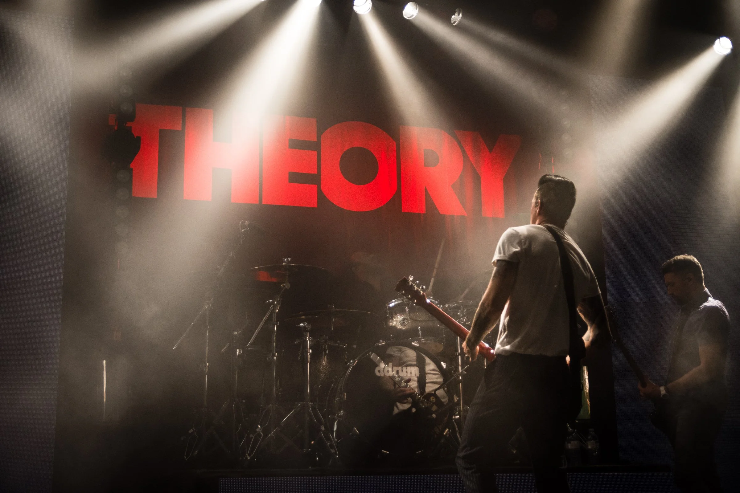 A stage with bright spotlights illuminating a band performing. A large red sign with the word 'THEORY' is displayed in the background. Two band members are visible, one with a guitar and the other with a bass guitar. The scene has a foggy or smoky at