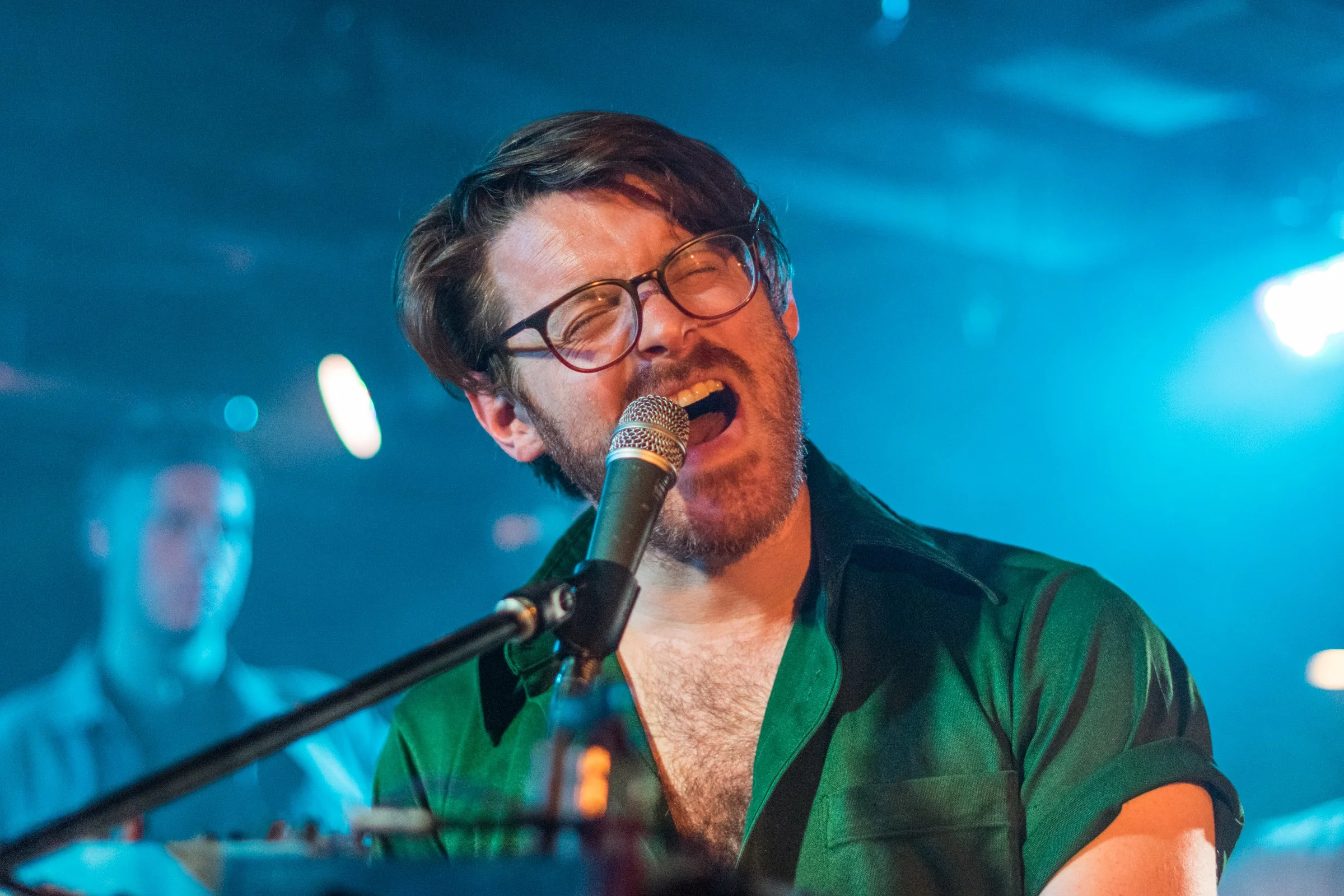 A man with glasses singing into a microphone on stage, smiling, with colorful stage lighting in the background.