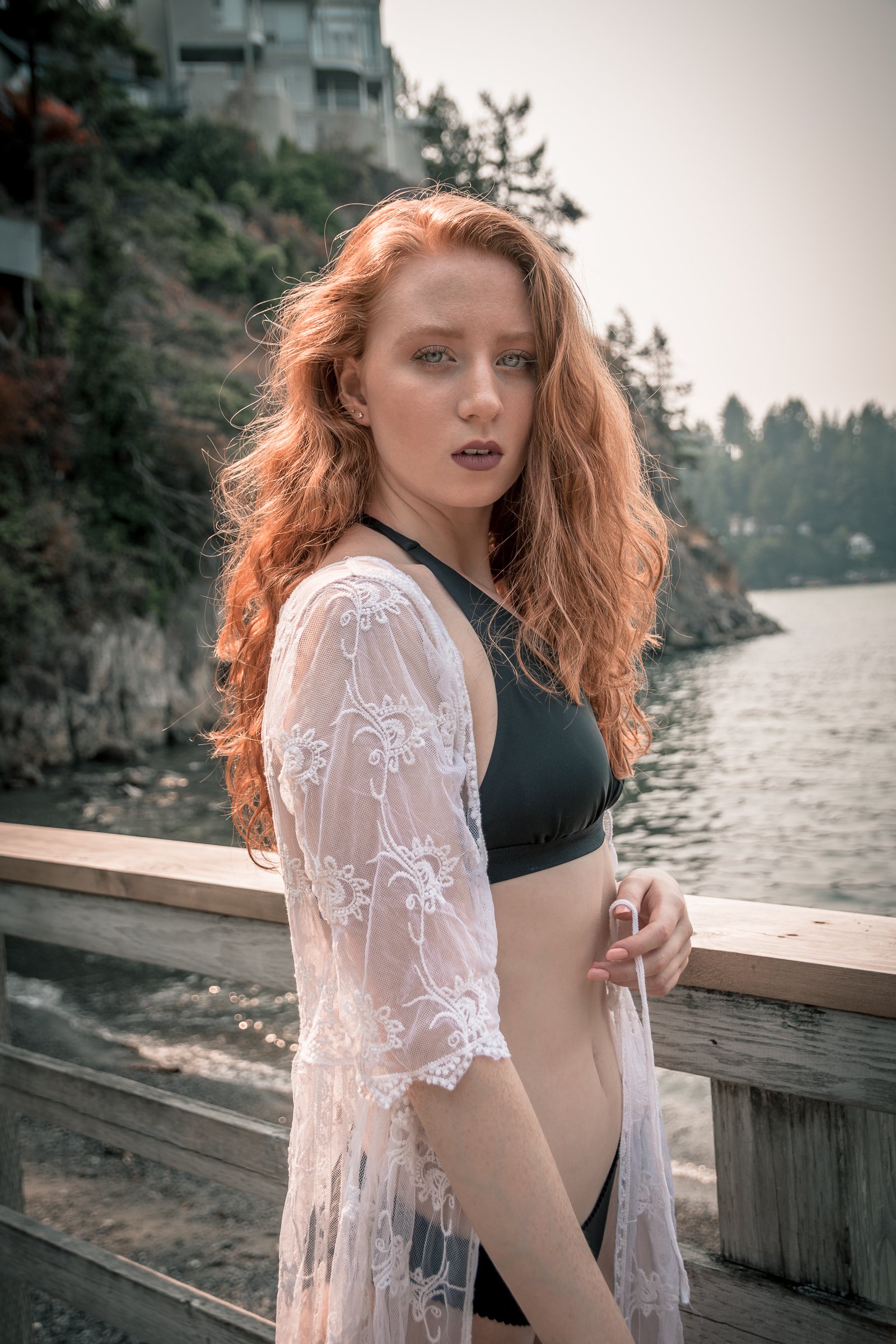 A young woman with long, wavy red hair and fair skin stands on a wooden dock near water, wearing a black bikini top and a sheer, white lace cover-up, with an overcast or sunset background with trees and houses on a hillside.
