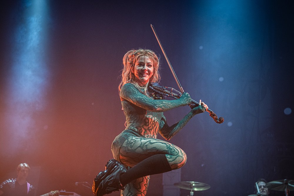 Performer dressed in a bodysuit with a snakeskin pattern playing a violin on stage, smiling.