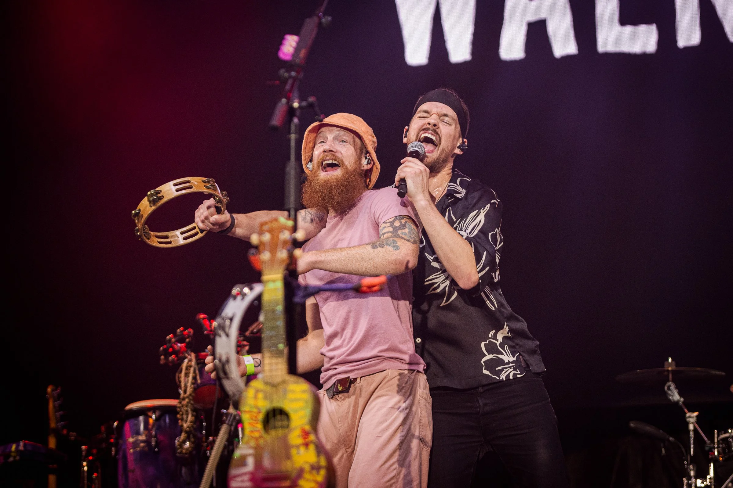 Two male performers are on stage singing passionately; one with a red beard and orange hat holding a tambourine, the other wearing a black headband and black patterned shirt holding a microphone. There are musical instruments in the foreground and a 
