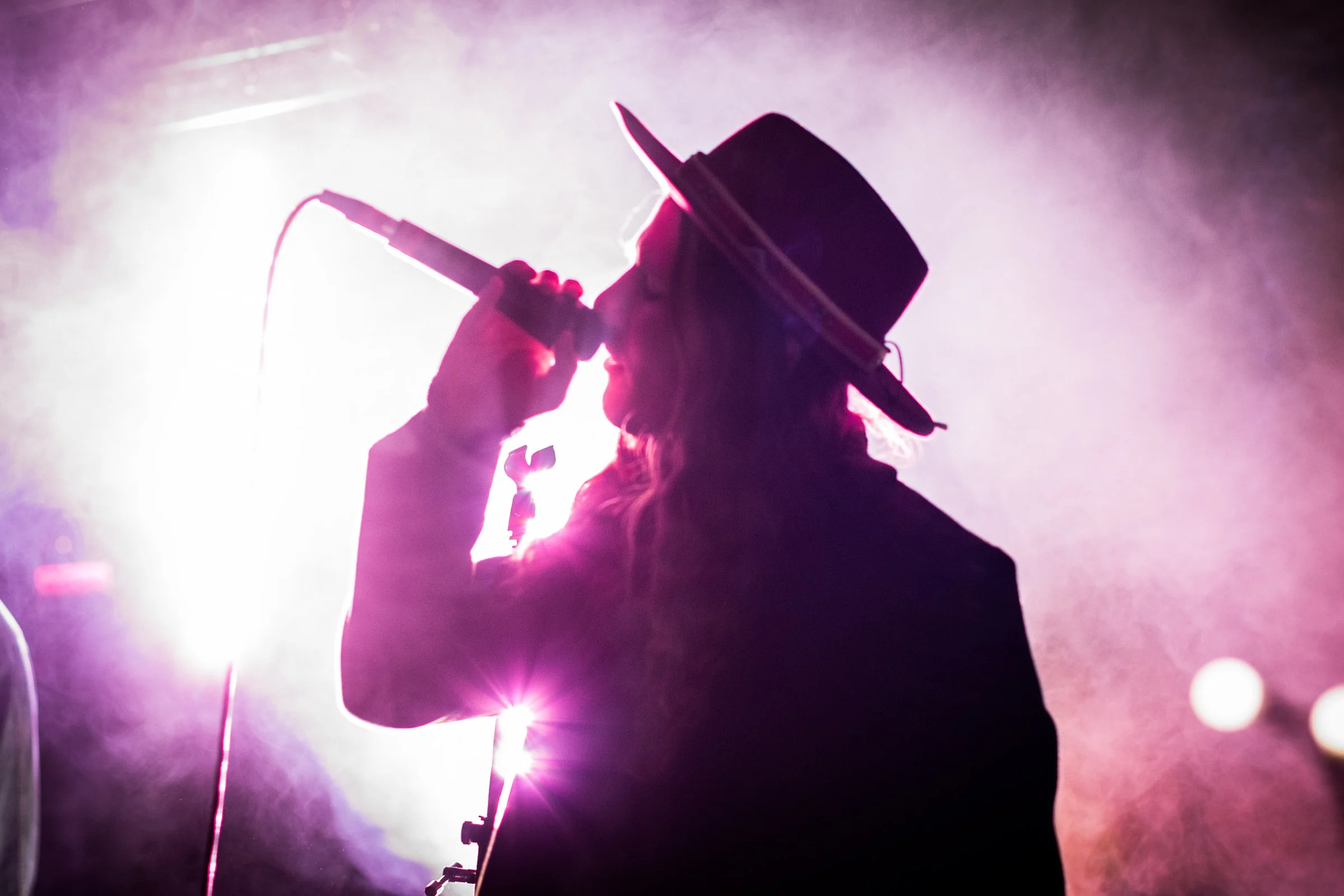 Silhouette of a person holding a microphone or similar object against bright stage lighting with pink and purple hues.