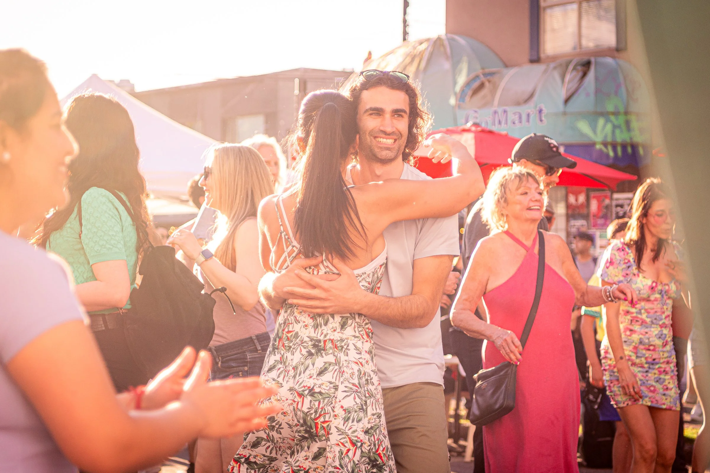 Man with long curly hair smiling and hugging woman at outdoor event with people walking and street tents