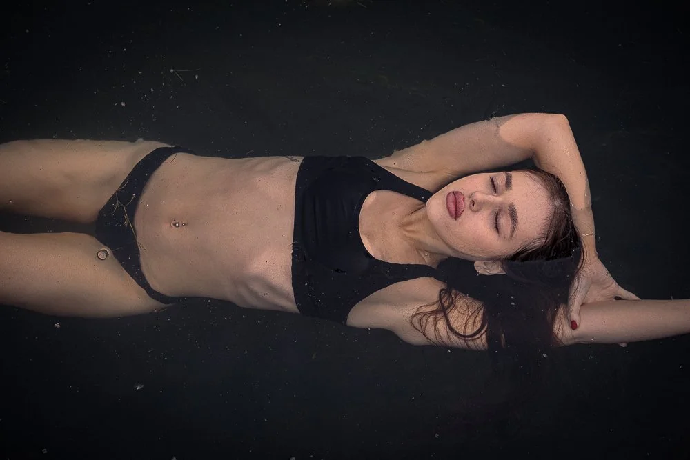 Woman lying in water with eyes closed, wearing a black sleeveless swimsuit, with long dark hair spread in water.
