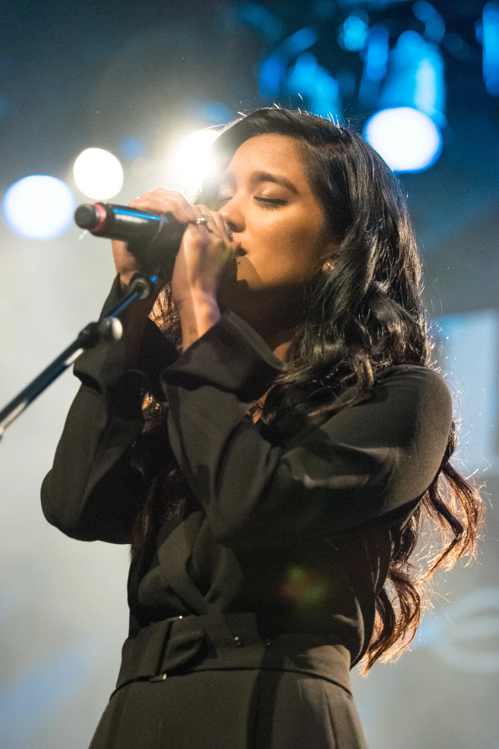 A woman with long dark hair singing into a microphone on stage, with bright lights and bokeh of blue lights in the background.