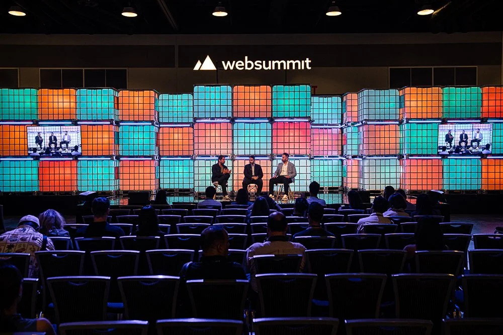 Panel discussion on stage at the Web Summit conference with three speakers, audience members seated, and a colorful illuminated backdrop with the Web Summit logo.