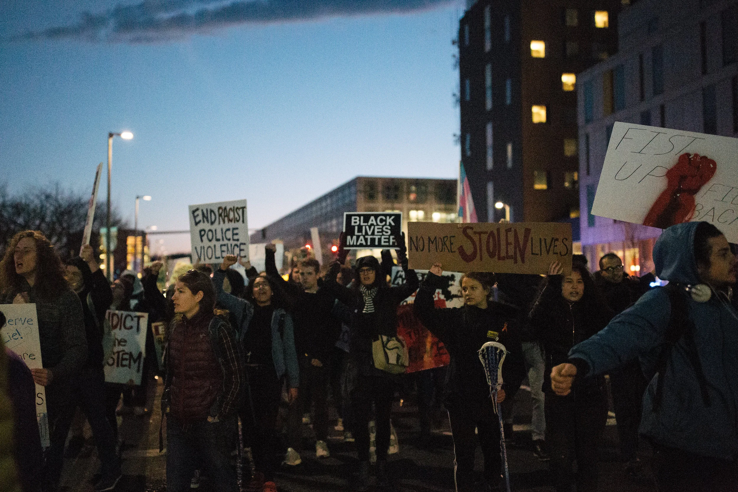 Photo description: Dozens of demonstrators stand facing the camera, mid-chant, some with their fists raised at the Not1More Rally Against Police Brutality hosted by MAAPB. Some hold signs saying ‘End Racist Police Violence’, ‘Black Lives Matter’, ‘No More Stolen Lives’, and ‘Fist Up Fight Back’. (Credit: Emily Goldhammer)