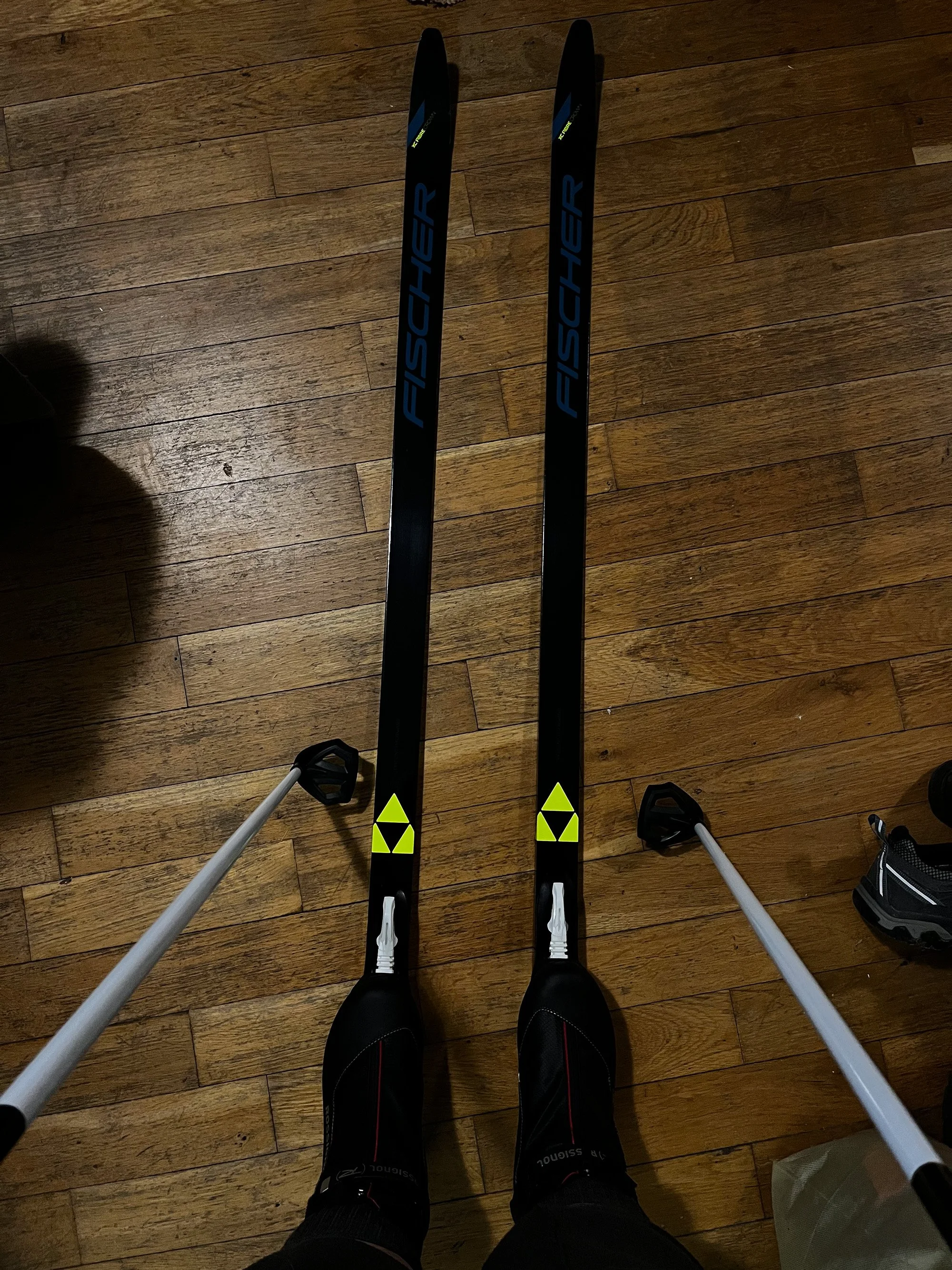 Point of view of a woman's feet in cross country boots and skis, standing with her ski poles on the wood floor of her living room.