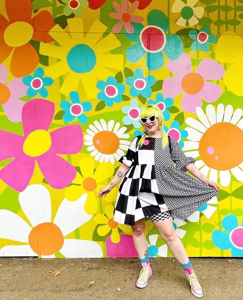 green haired dompaine dressed young woman with black and white checkerd dress standing infront of dressew happy floral mural .jpg